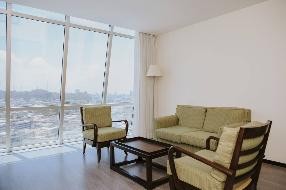 Een moderne hotelkamer met een groot raam dat uitzicht geeft op de stad, een groen gestreepte bank, twee fauteuils en een kleine houten salontafel.