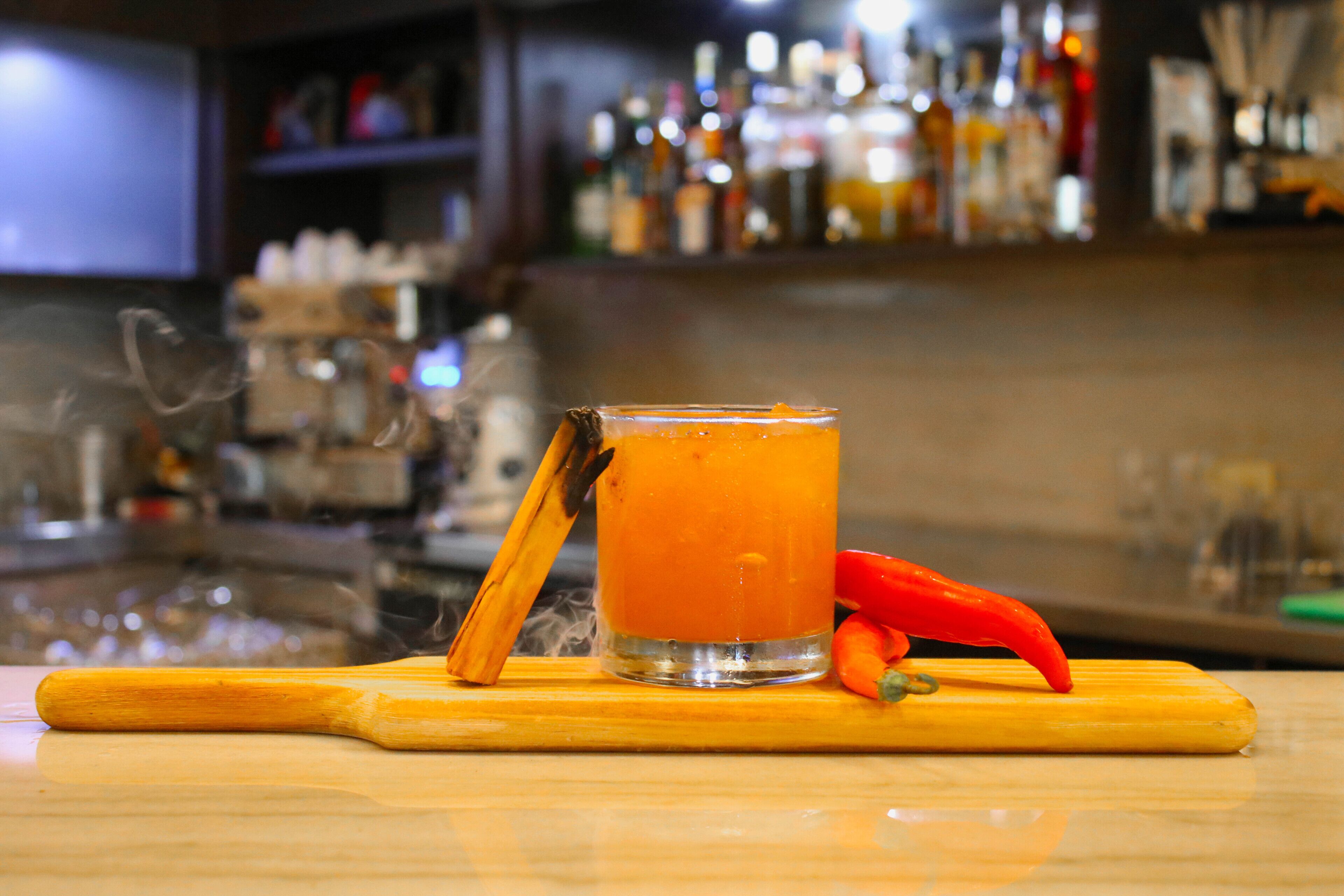 A cocktail glass with a cinnamon stick on a wooden bar mat, with a blurred background of a bar setting.