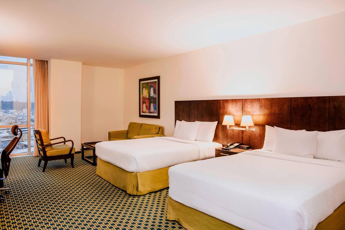 Une chambre d’hôtel avec deux lits, un bureau, une chaise, une fenêtre donnant sur la ville et un tableau au mur.