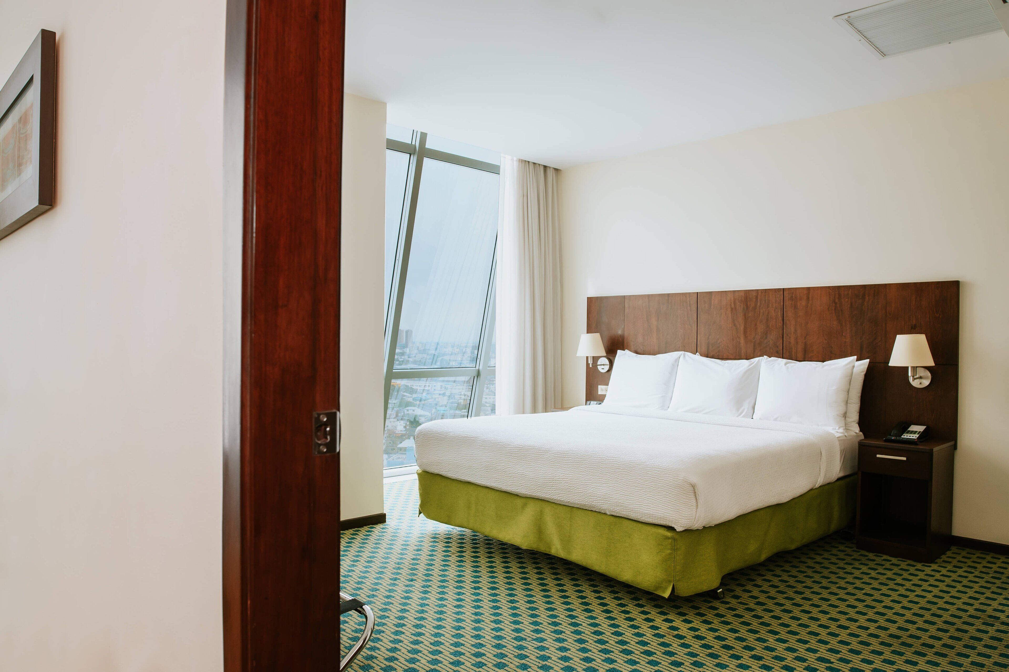 Un cuarto de hotel con una cama grande, dos lámparas de noche, una mesita de noche y vistas a la ciudad a través de un ventanal amplio.