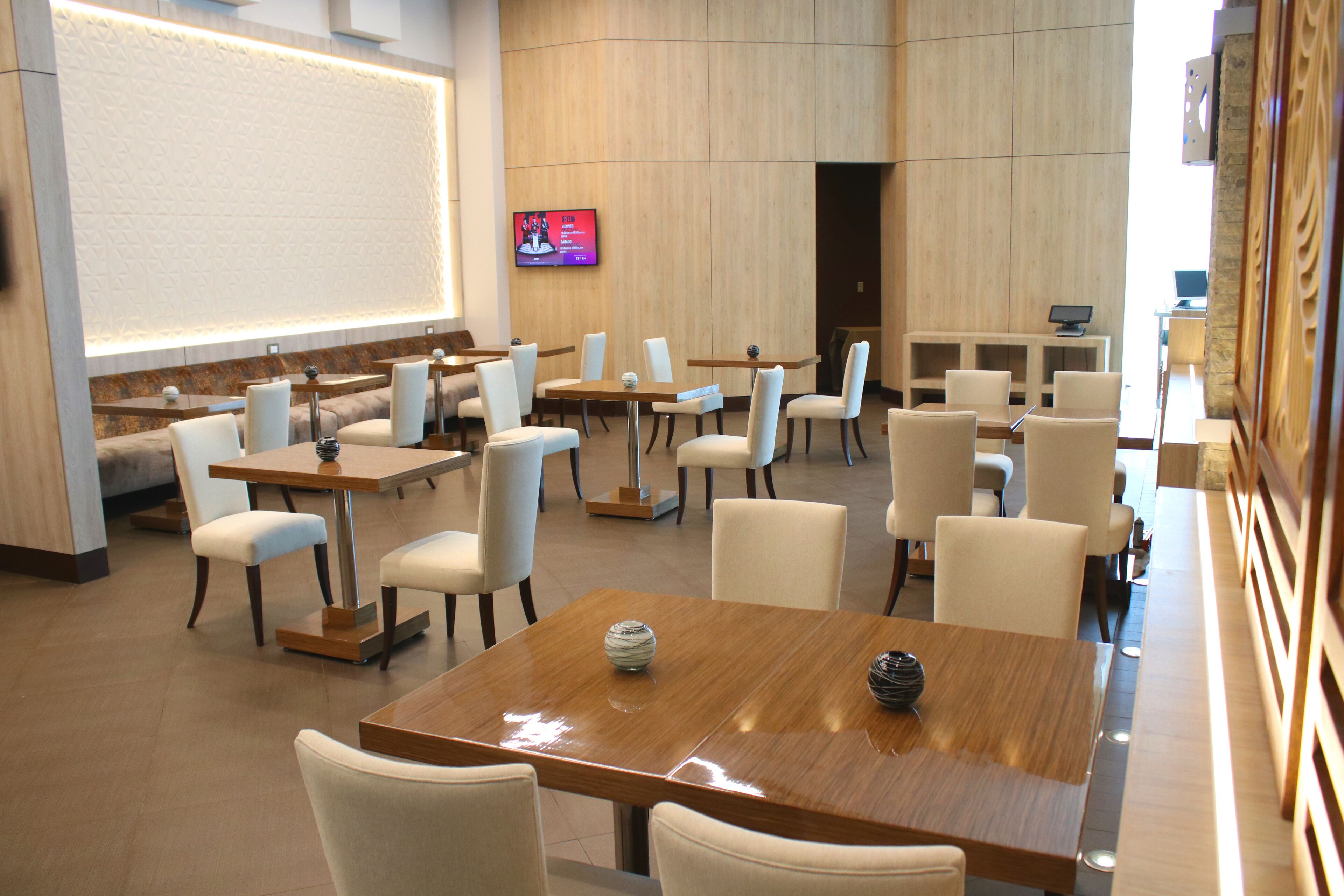 Eine moderne Hotellobby mit mehreren Sitzbereichen, darunter Tische und Stühle, und einem großen Bildschirm, der Inhalte anzeigt.