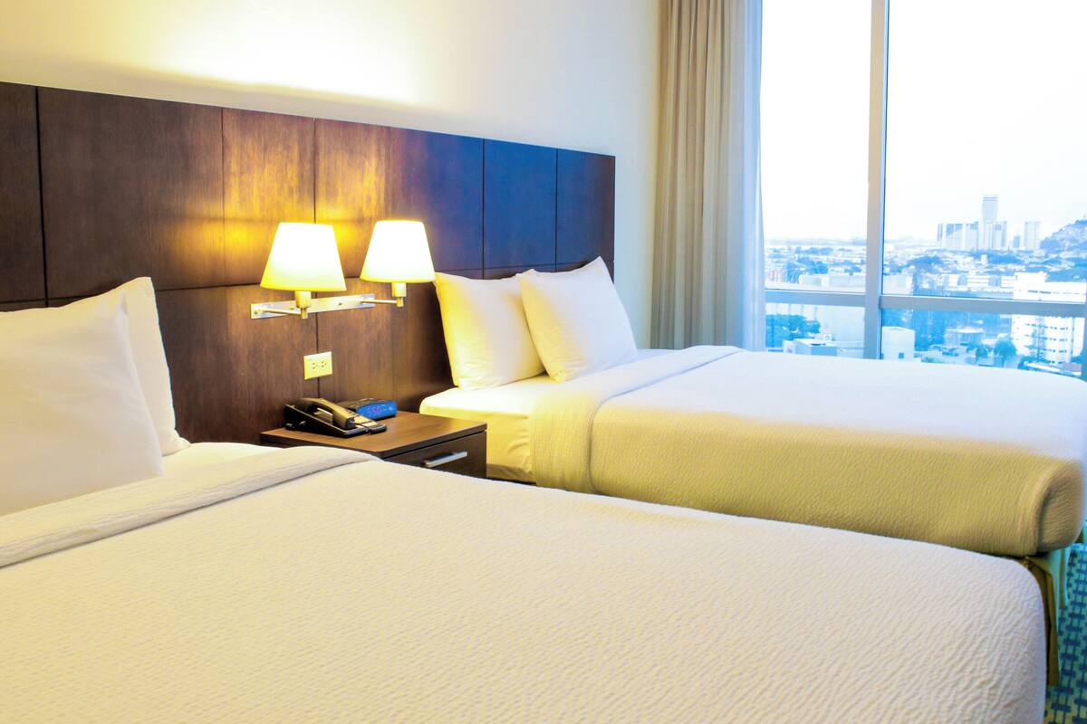 Een hotelkamer met twee bedden, een houten hoofdbord, nachtlampjes en uitzicht op de stad door grote ramen.
