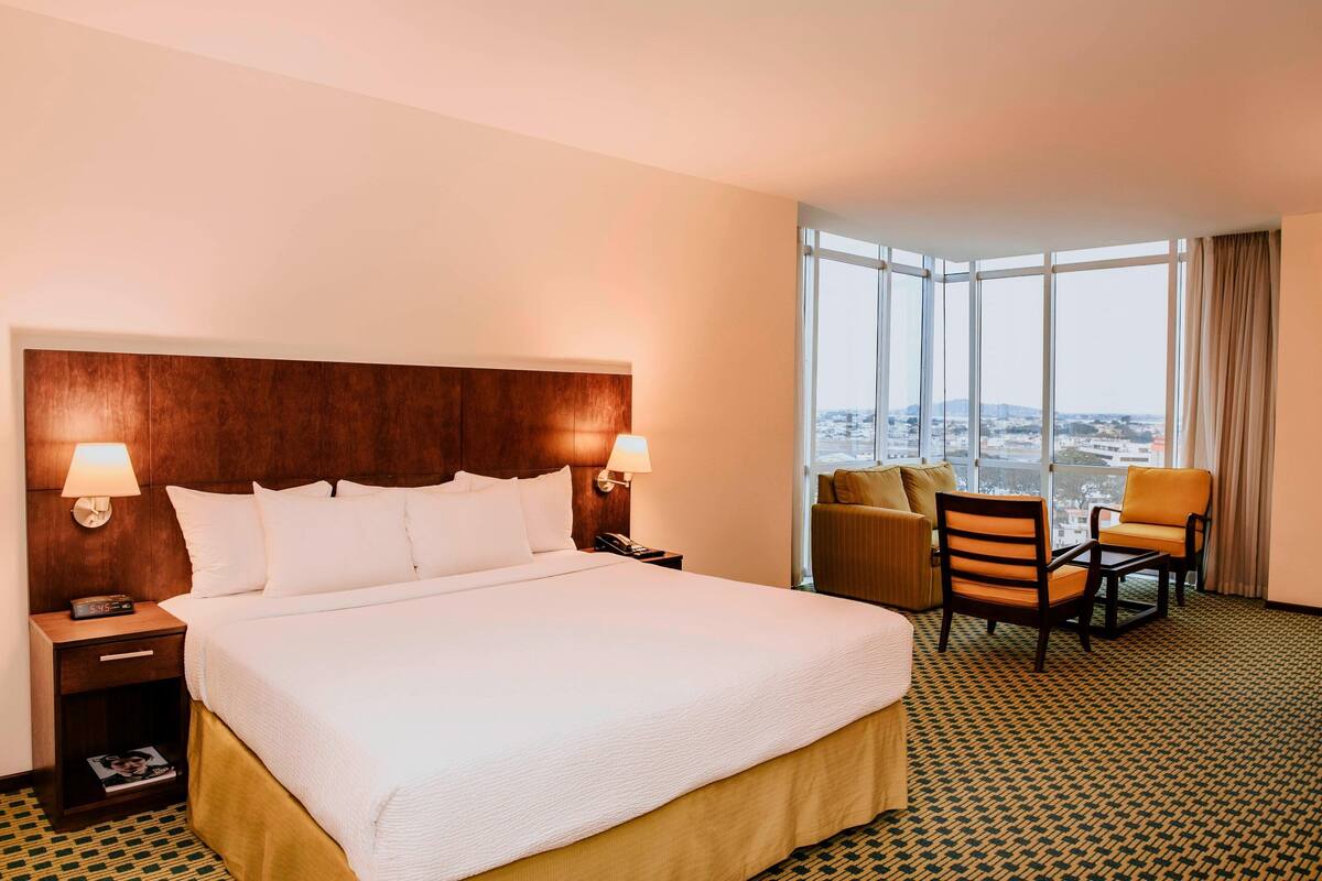 Une chambre d’hôtel dotée d’un grand lit, de deux tables de chevet avec des lampes, d’une vue sur la ville grâce à de grandes fenêtres et d’un coin salon avec deux fauteuils.