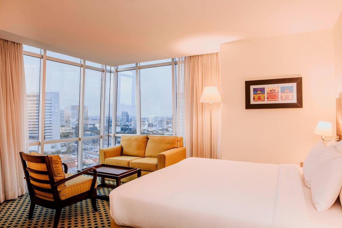 Une chambre d’hôtel avec un grand lit, un canapé, un fauteuil, une petite table et une vue sur la ville grâce à de grandes fenêtres.