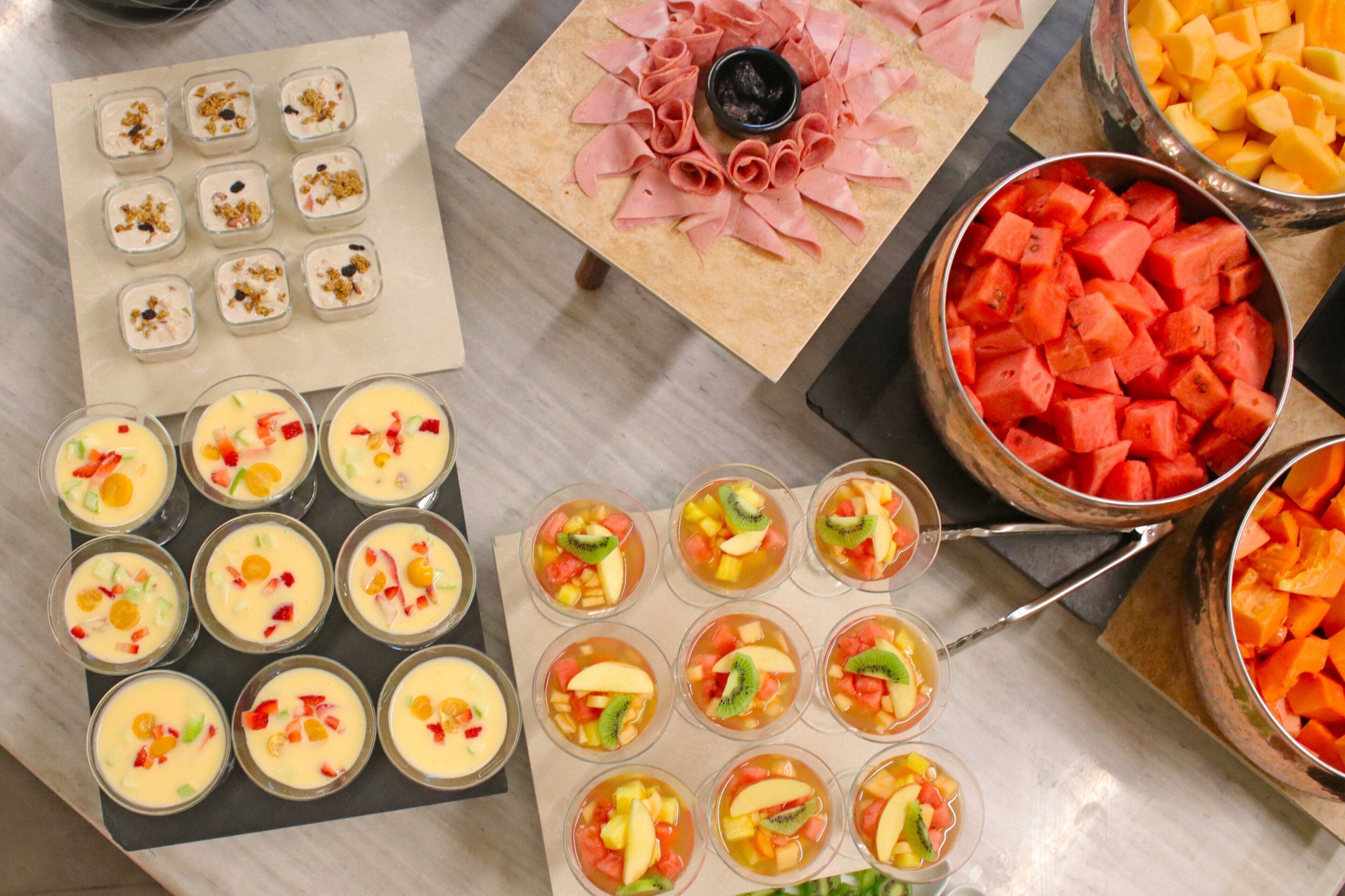 A buffet table with various food items including fruit cups, sliced meats, and bowls of fruit.