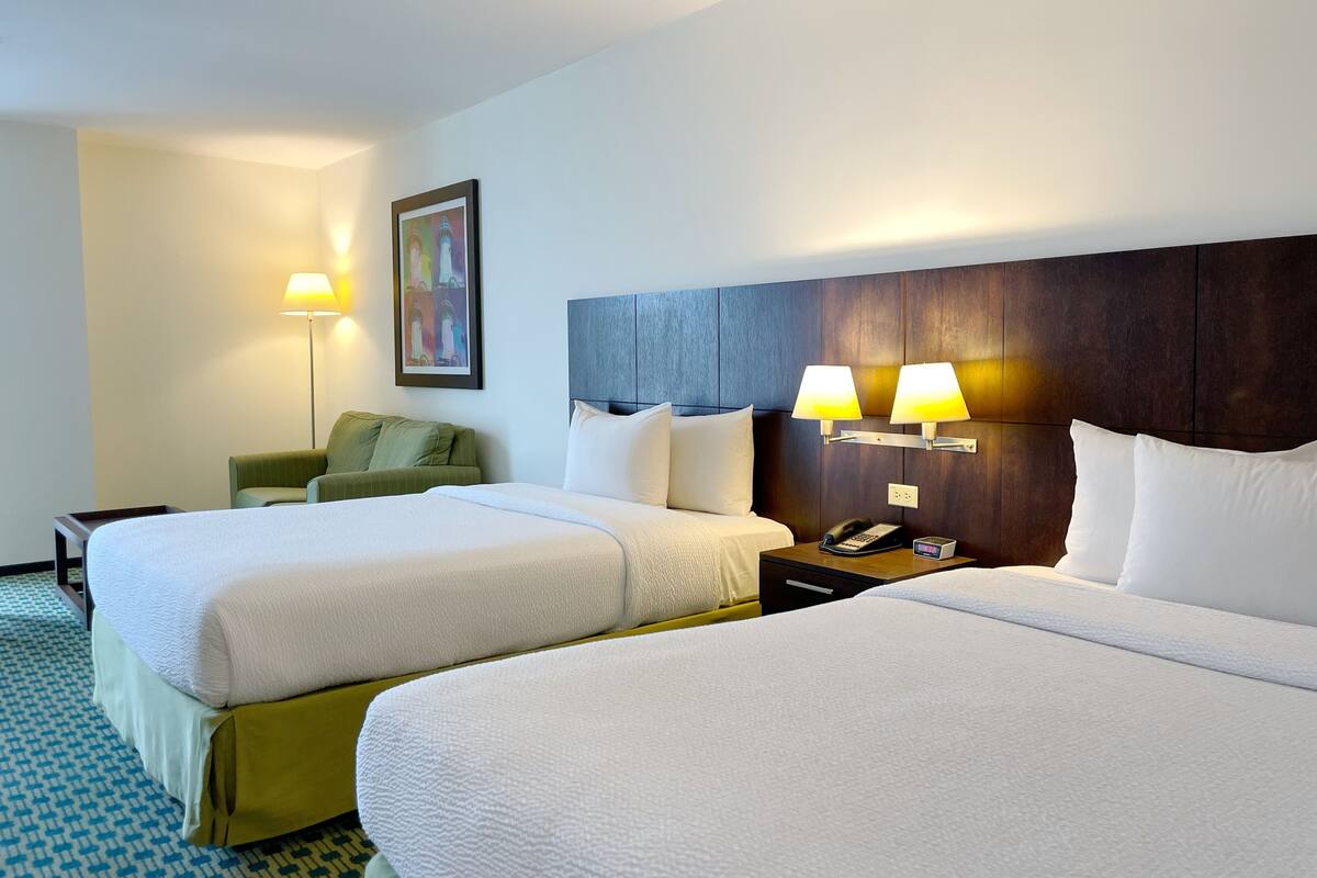 Een hotelkamer met twee bedden, een nachtkastje met telefoon en digitale klok, een groene fauteuil, een lamp en een schilderij aan de muur.
