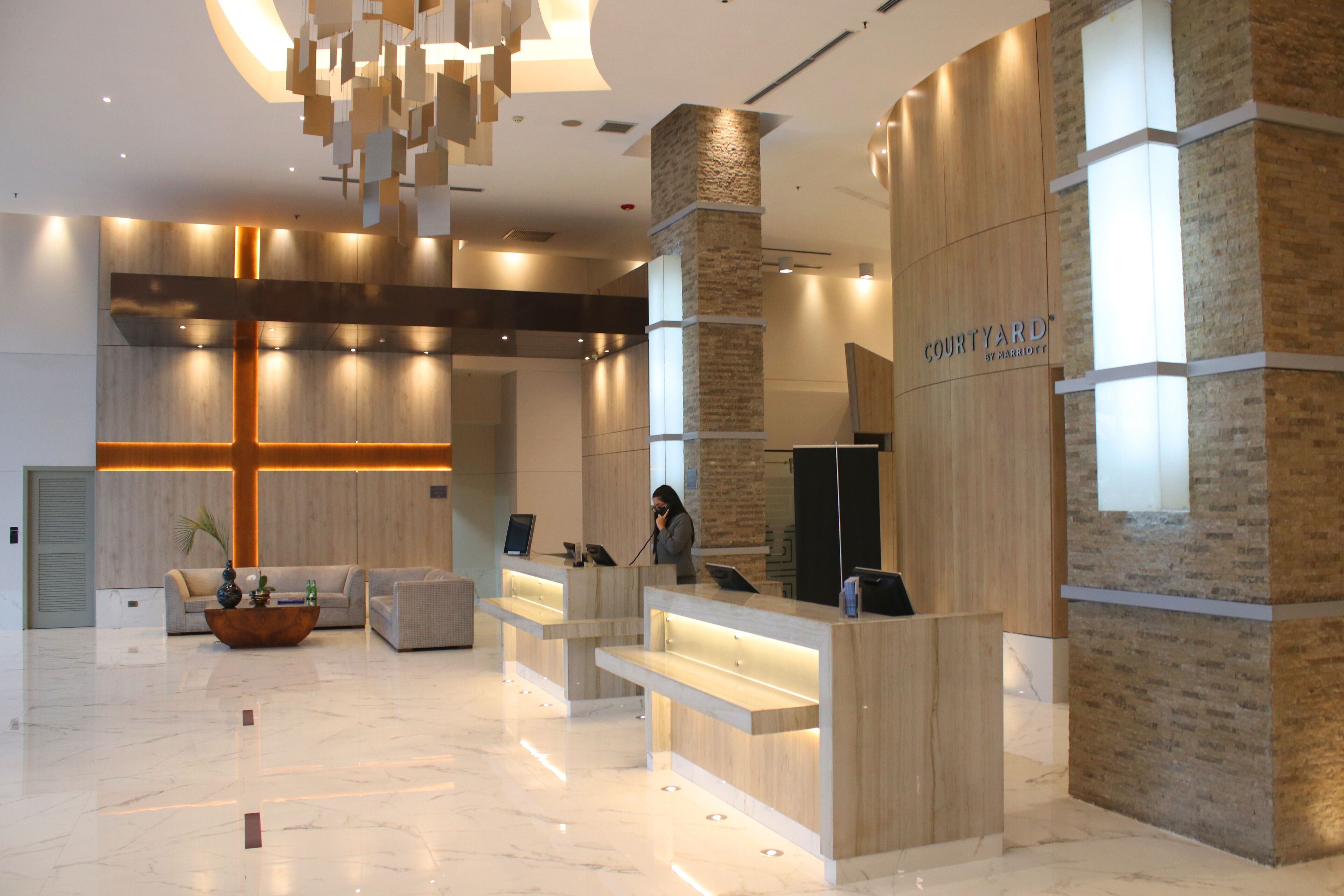 Un hall d’hôtel moderne avec un comptoir d’accueil, un espace salon et un grand luminaire en forme de croix au plafond.