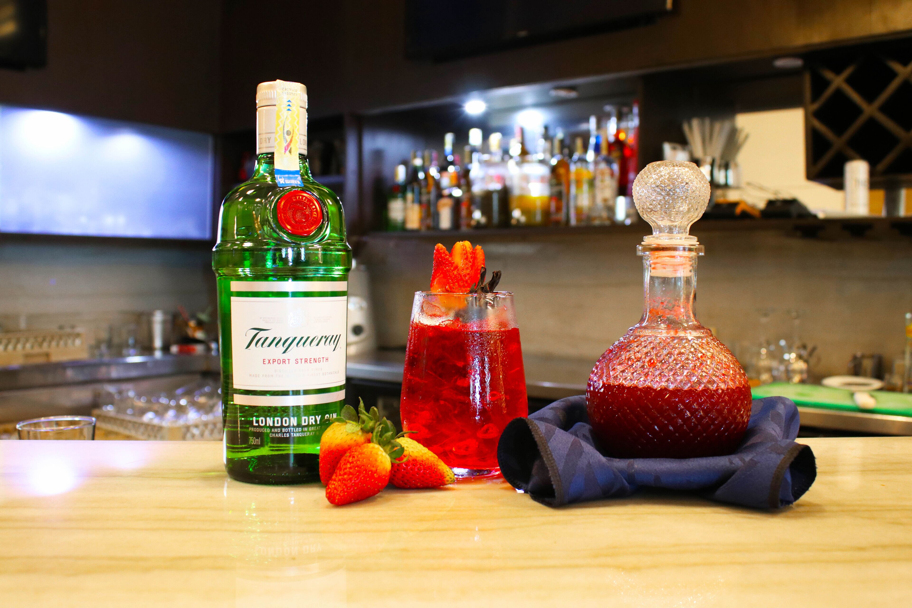 Un bar con una botella de ginebra London Dry, un cóctel adornado con fresa y una botella de jarabe rojo.