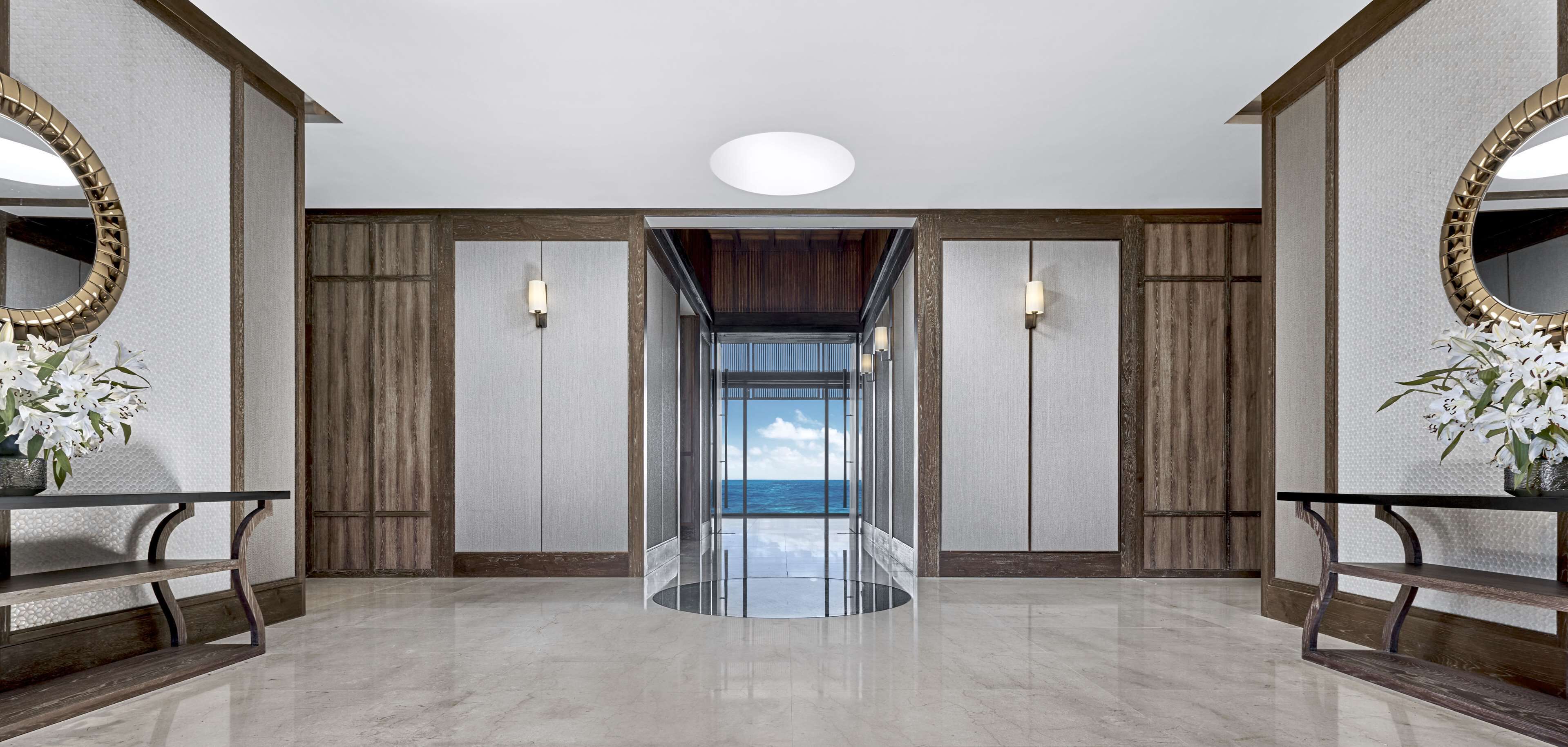 A modern hotel lobby with a marble floor, wooden panel walls, and a glass elevator shaft leading to a balcony with a view of the sea.