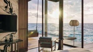 A modern room with a large window overlooking the sea, a flat-screen TV, a wooden bench, and a lamp. - Park Hyatt Maldives Hadahaa (Hadahaa)