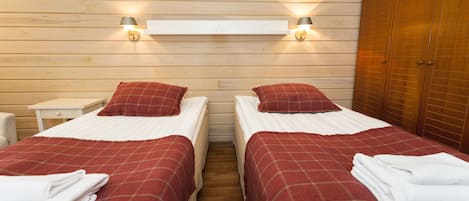 Habitación doble con dos camas, cada una con colcha a cuadros rojos y almohadas blancas. Hay dos mesitas de noche con lámparas, un armario de madera y un estante blanco sobre las camas.