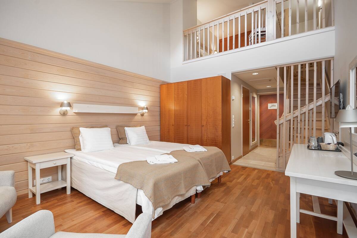 A modern hotel room with a wooden floor, a large bed with two pillows, a bedside table, a wardrobe, and a staircase leading to an upper level.