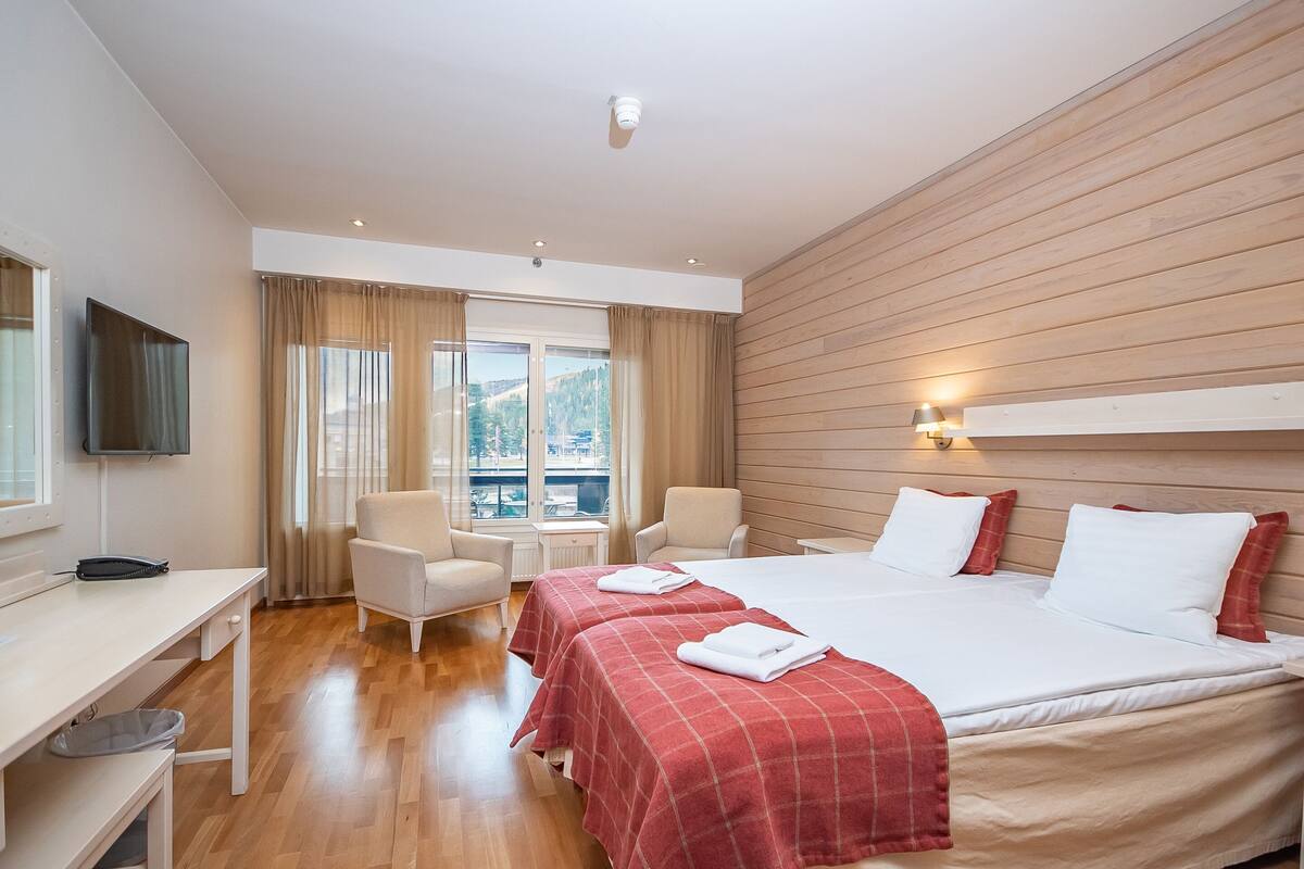 A hotel room with a bed, two bedside tables, a desk, a chair, a TV, and a window with curtains.