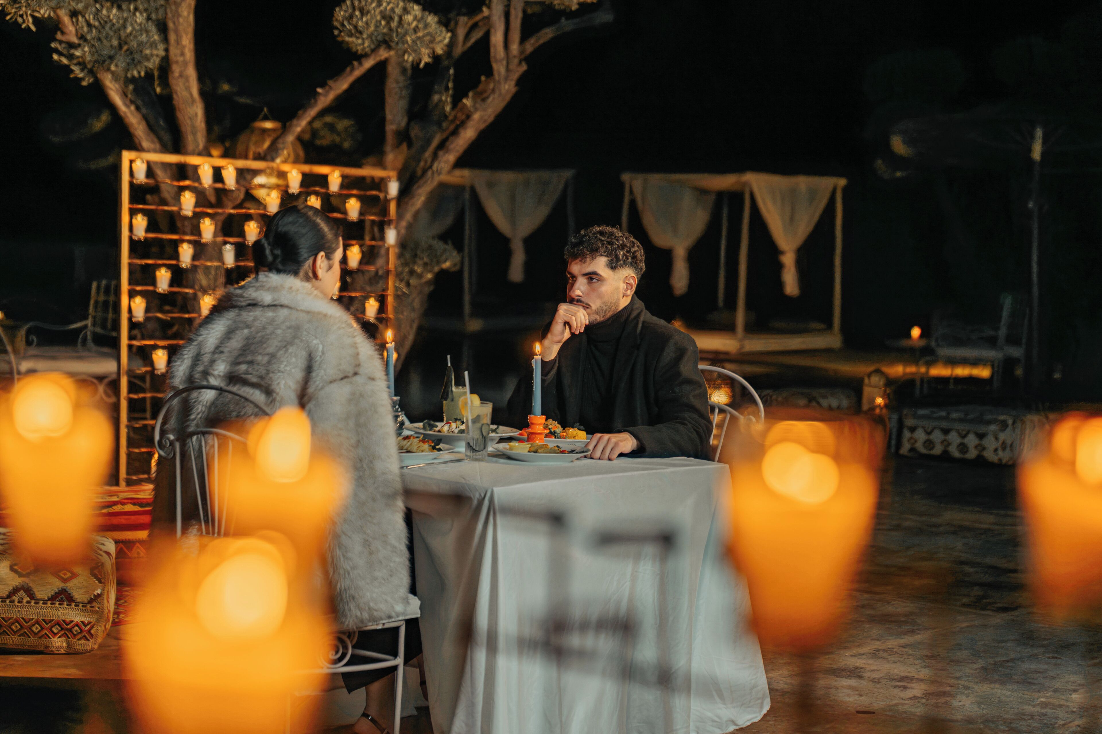 Dos personas sentadas en una mesa al aire libre con mantel blanco, rodeadas de árboles y una estructura decorativa de luces.