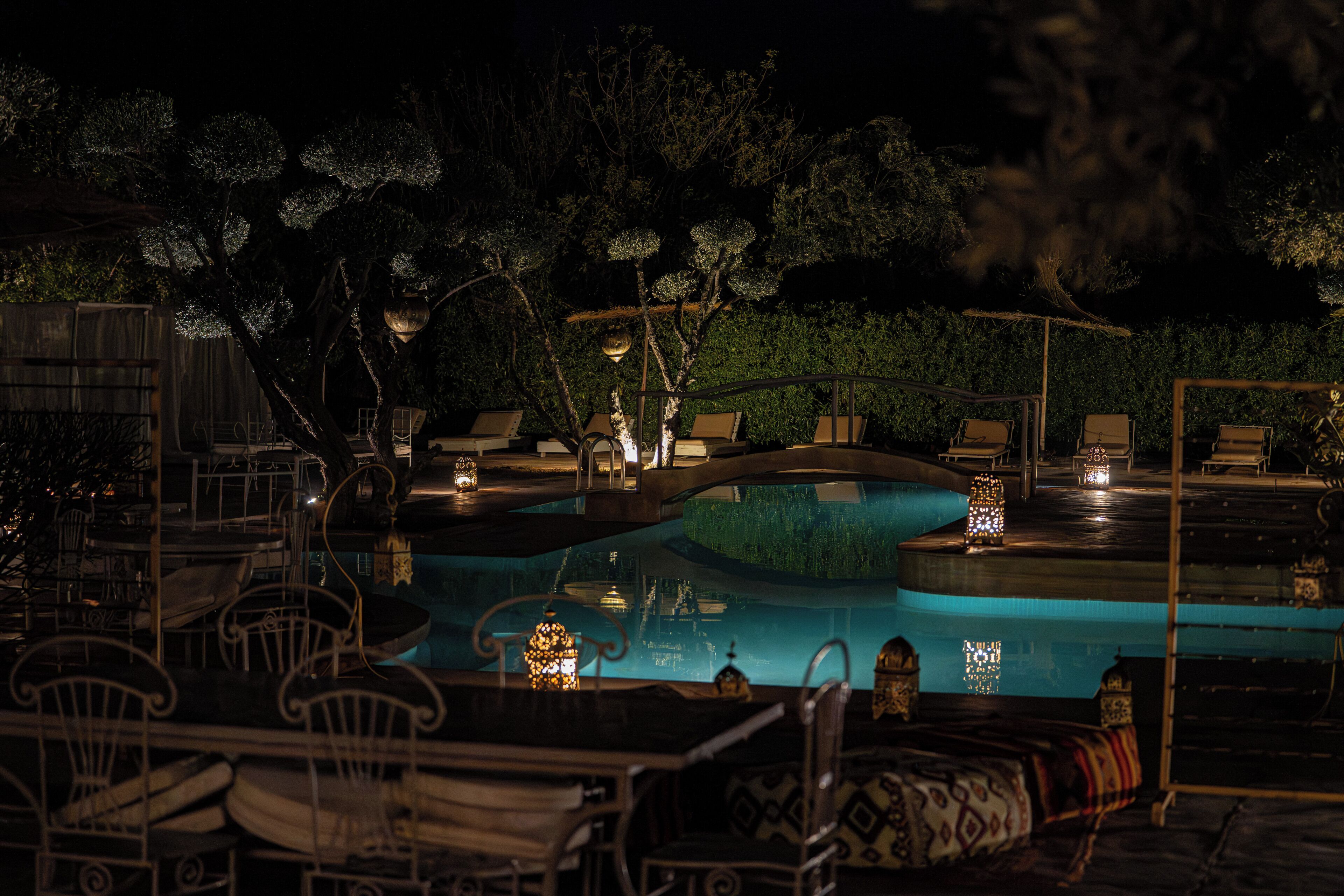 Zona de piscina exterior con iluminación nocturna, sillones reclinables y mesas, rodeada de árboles y una valla de madera.
