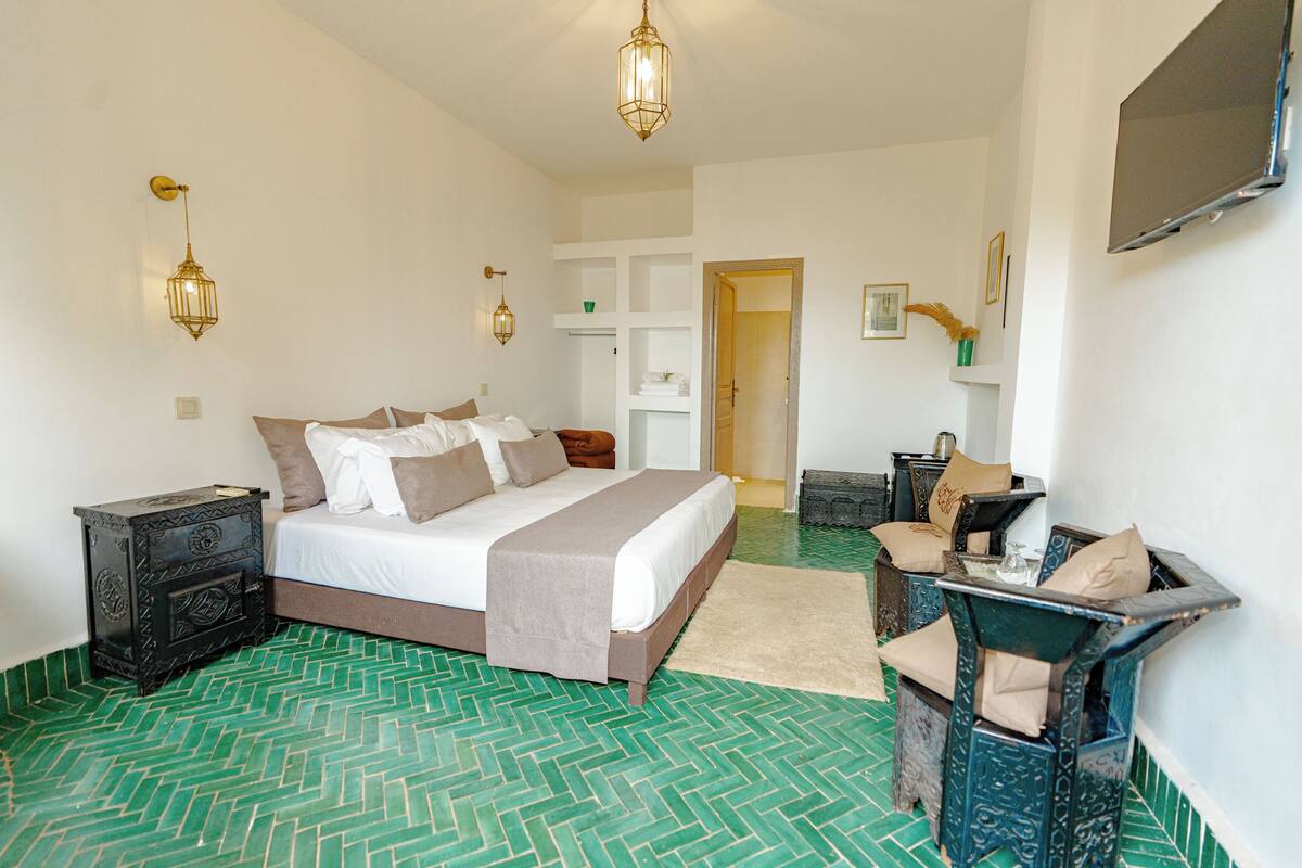 A bedroom with a bed, bedside tables, a chair, a TV, and a door leading to another room.
