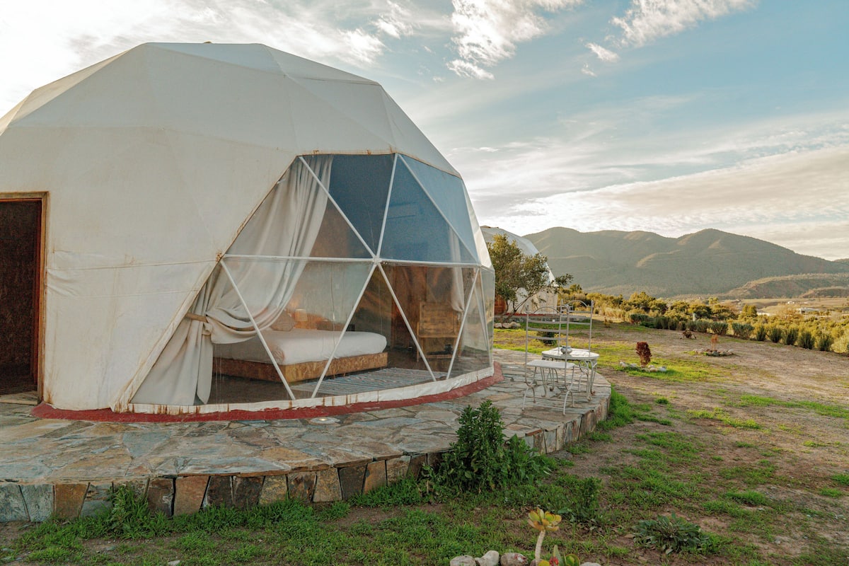 A geodesic dome structure with a bed and a small table, surrounded by a stone patio and a grassy area with mountains in the background.