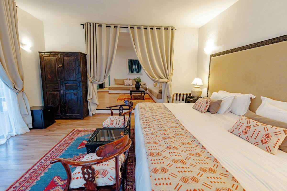 A hotel room with a large bed, a wooden wardrobe, a sofa, a small table, and a chair.