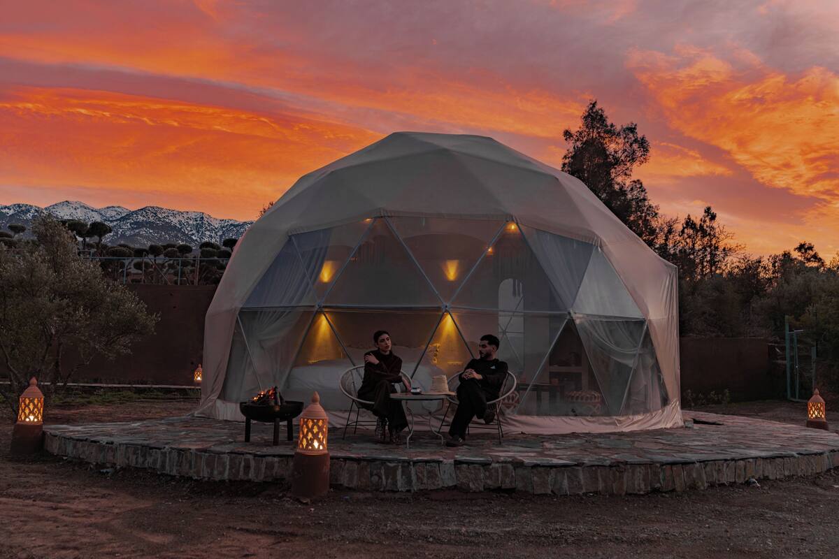A geodesic dome tent with two people seated at a table, surrounded by lanterns and a stone platform.