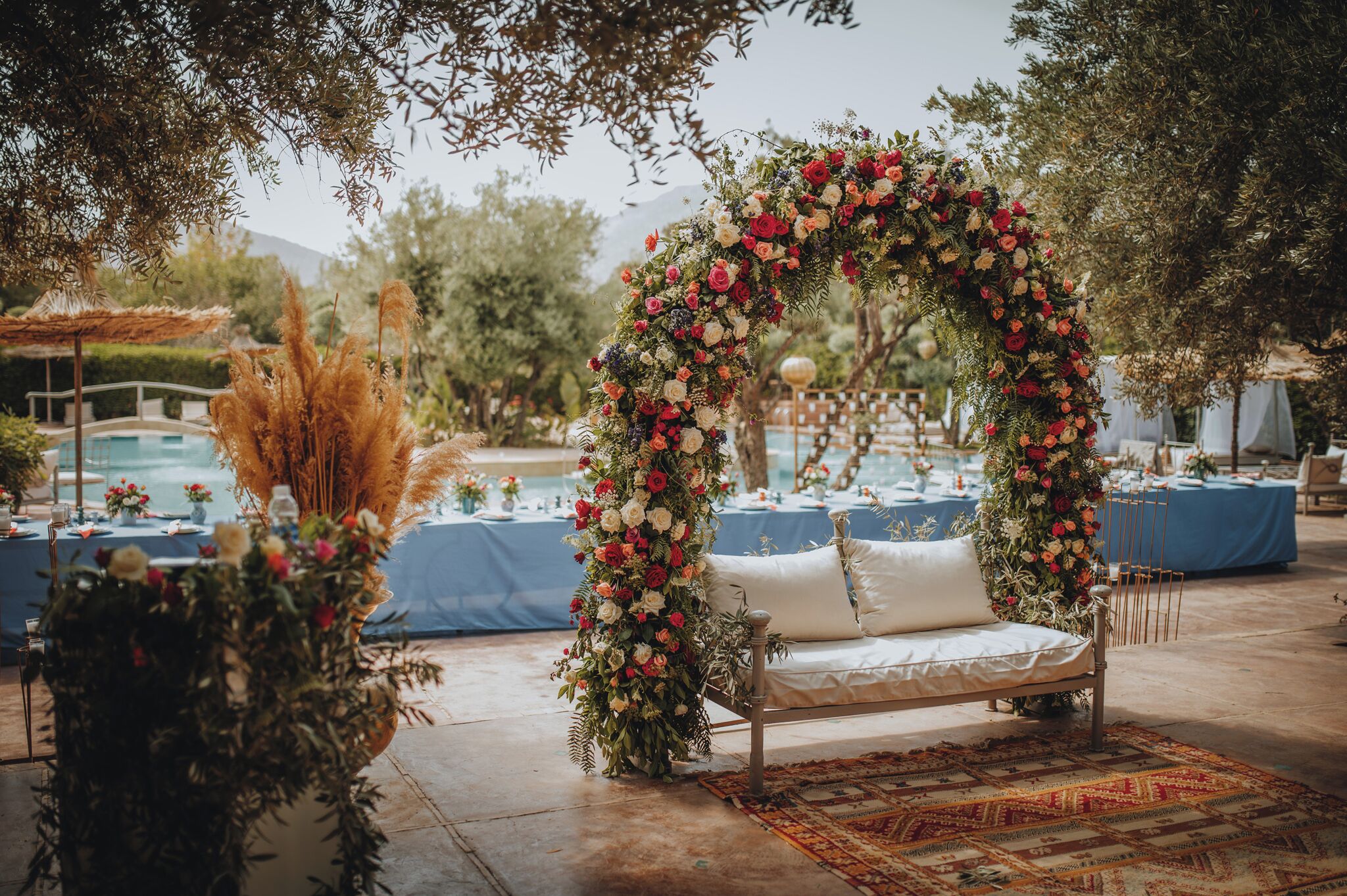 Montaje de boda al aire libre con un arco floral, un sofá blanco y mesas con manteles azules.