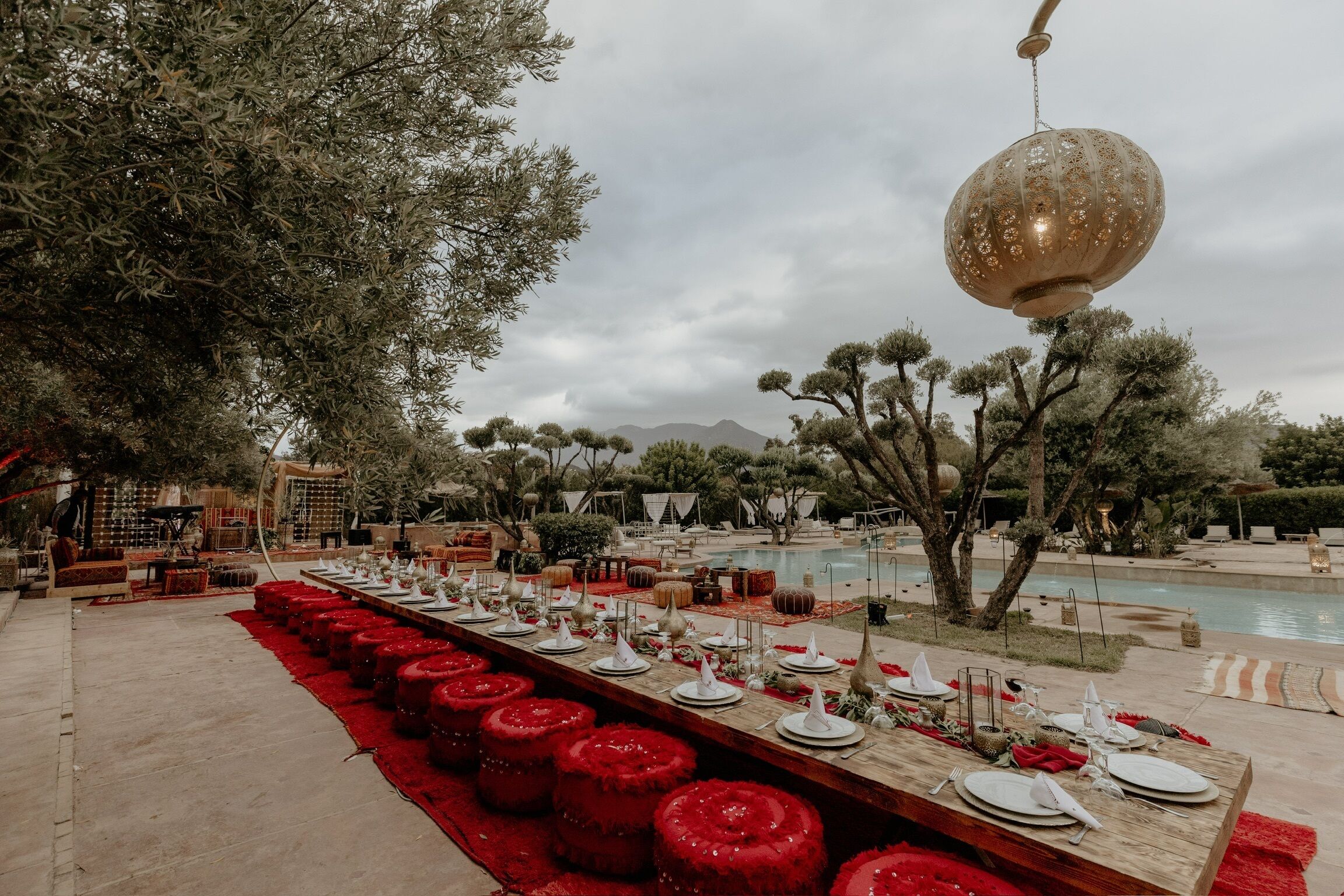A long dining table set with white plates and red accents, surrounded by trees and a pool in a resort setting.