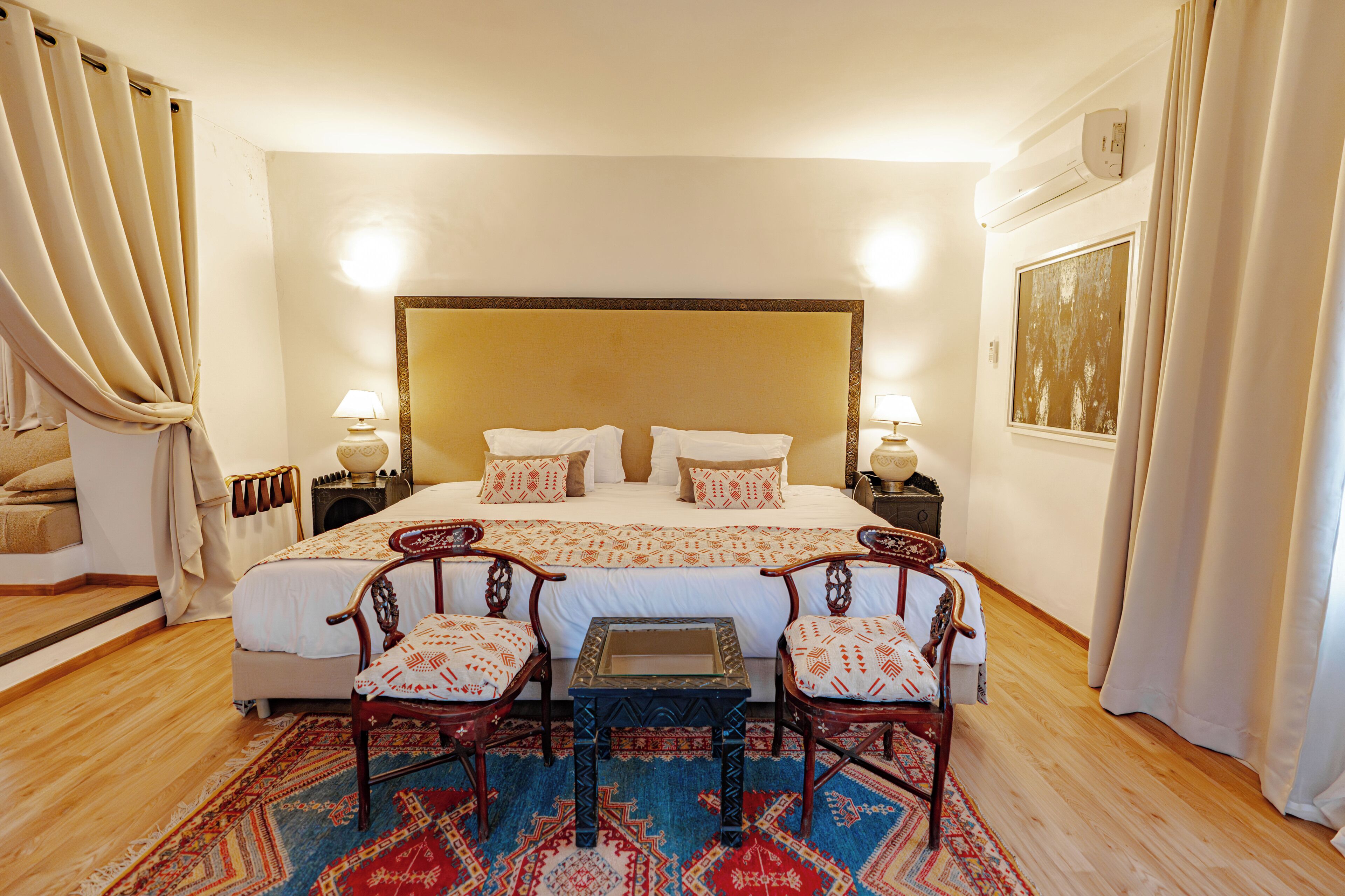 A hotel room with a large bed, two bedside tables, a small table, four chairs, a patterned rug, and a window with curtains.