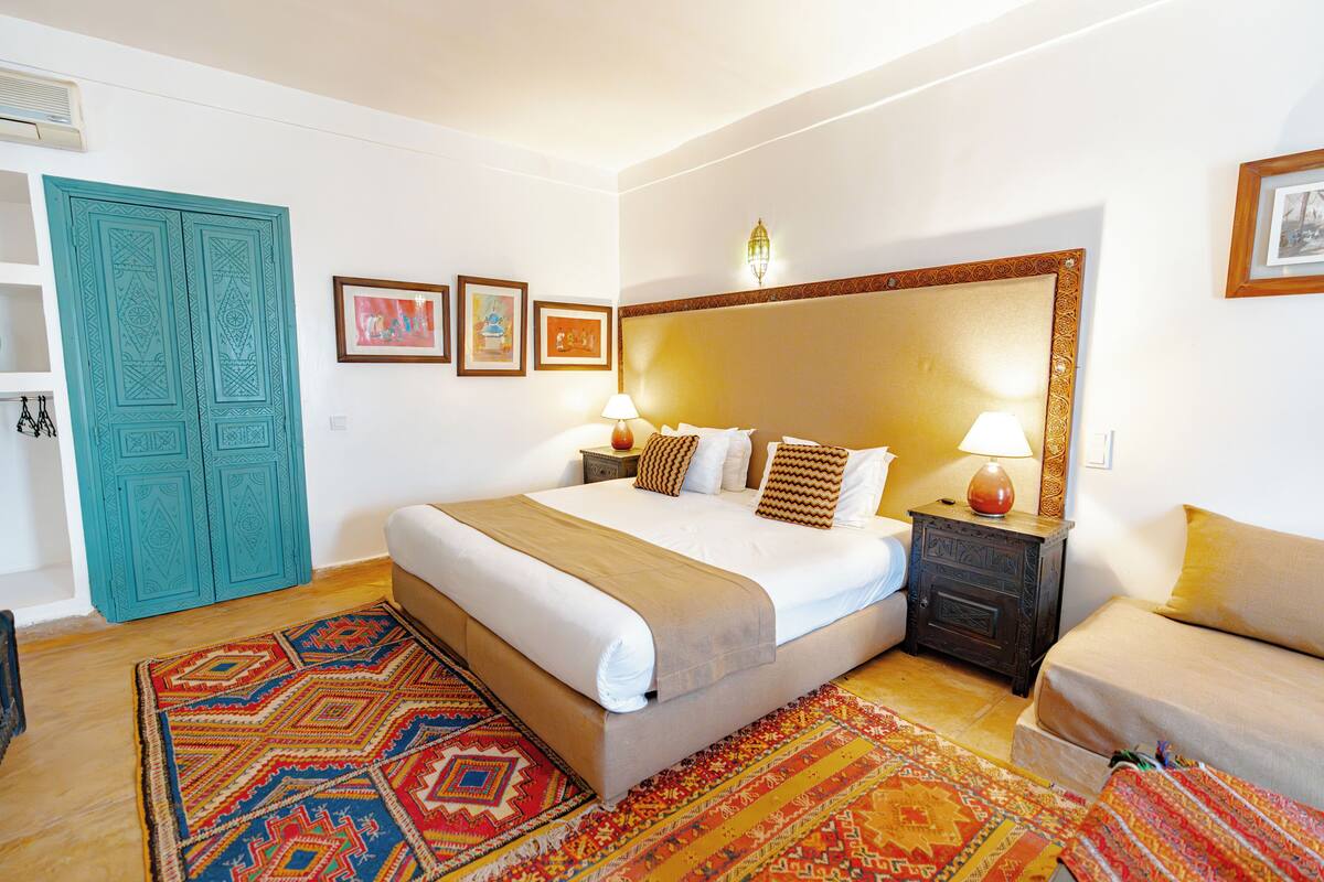 A bedroom with a large bed, a nightstand, a dresser, a sofa, a patterned rug, and a teal door.