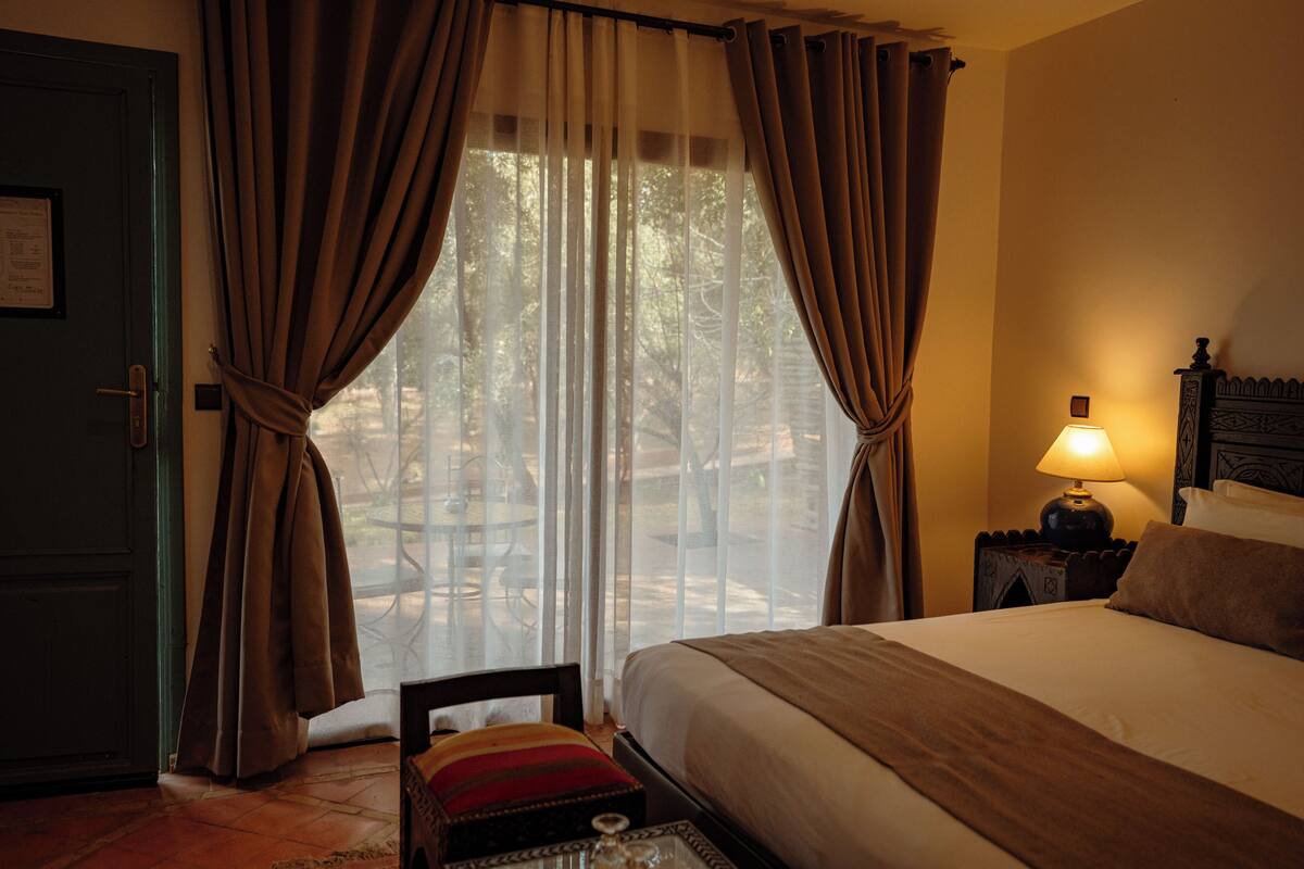A hotel room with a bed, curtains, a nightstand with a lamp, and a view through the window.