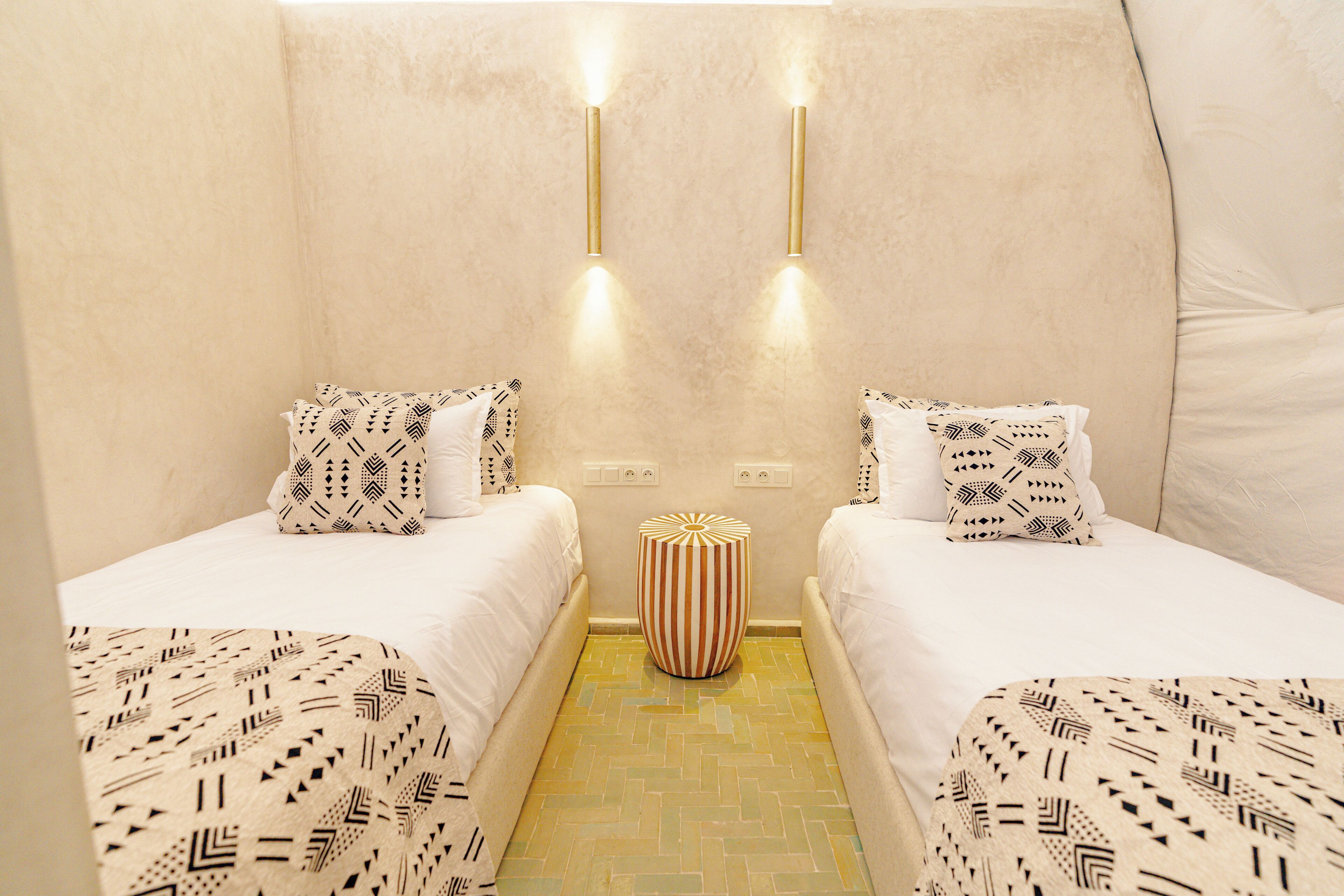 A hotel room with two beds, patterned bedding, a striped ottoman, and wall-mounted lights.