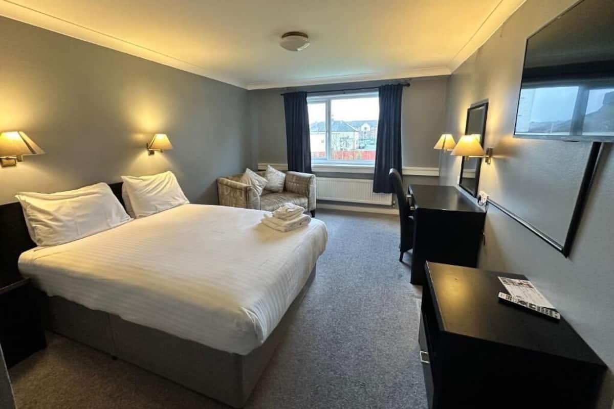A hotel room with a bed, bedside tables, a desk, a chair, a TV, and a window with curtains.