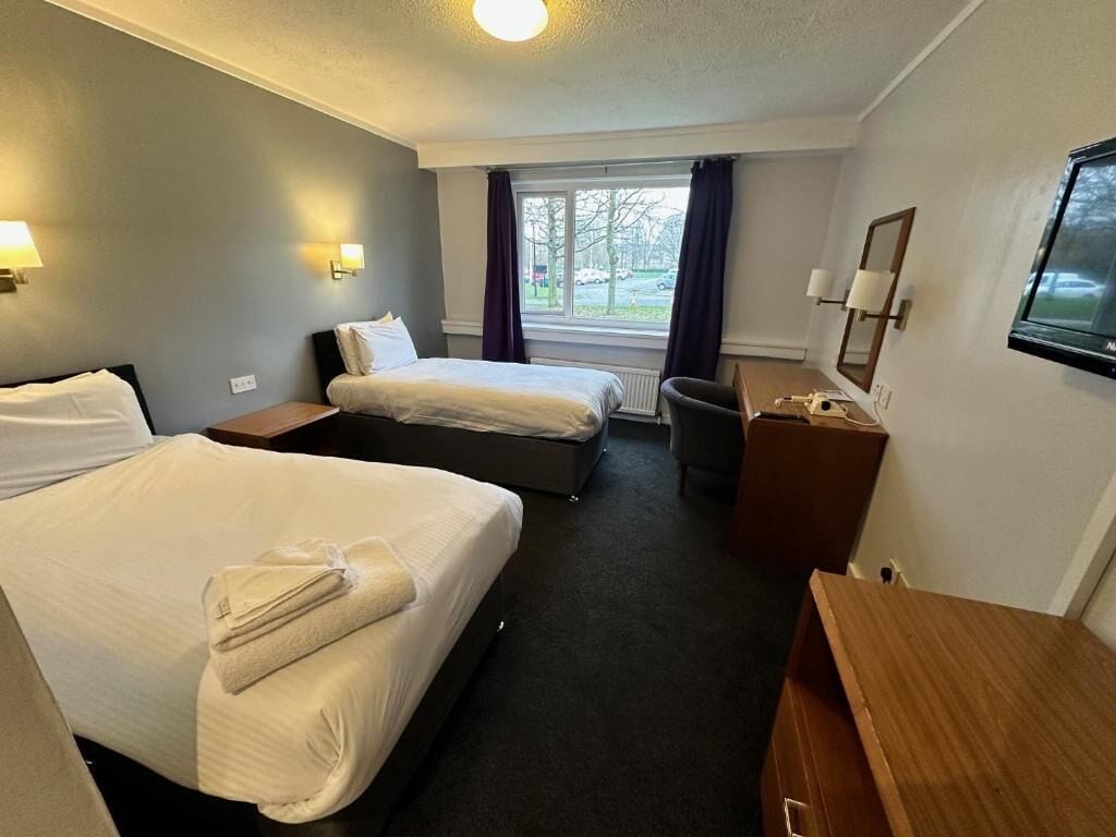 A hotel room with two beds, a desk, a chair, a television, and a window with curtains.