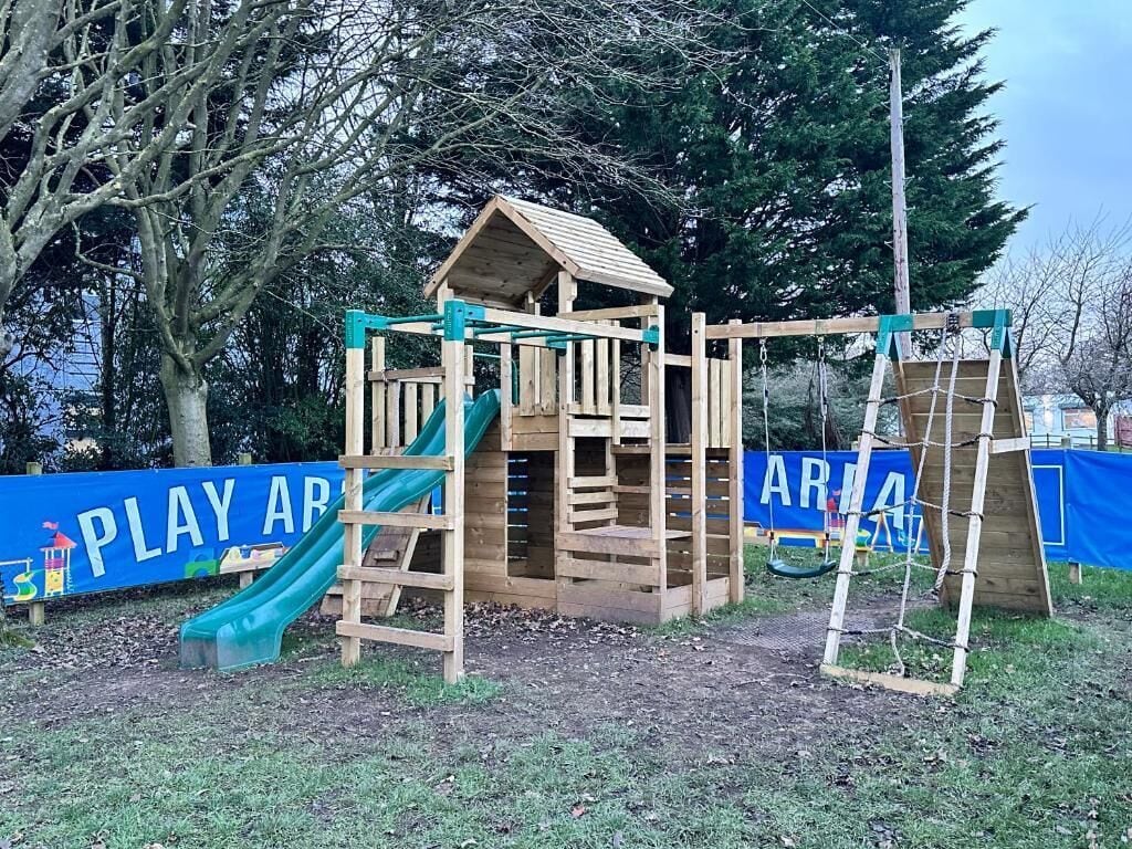A wooden play structure with a slide and climbing area, surrounded by trees and a blue banner with 'PLAY AREA' text.