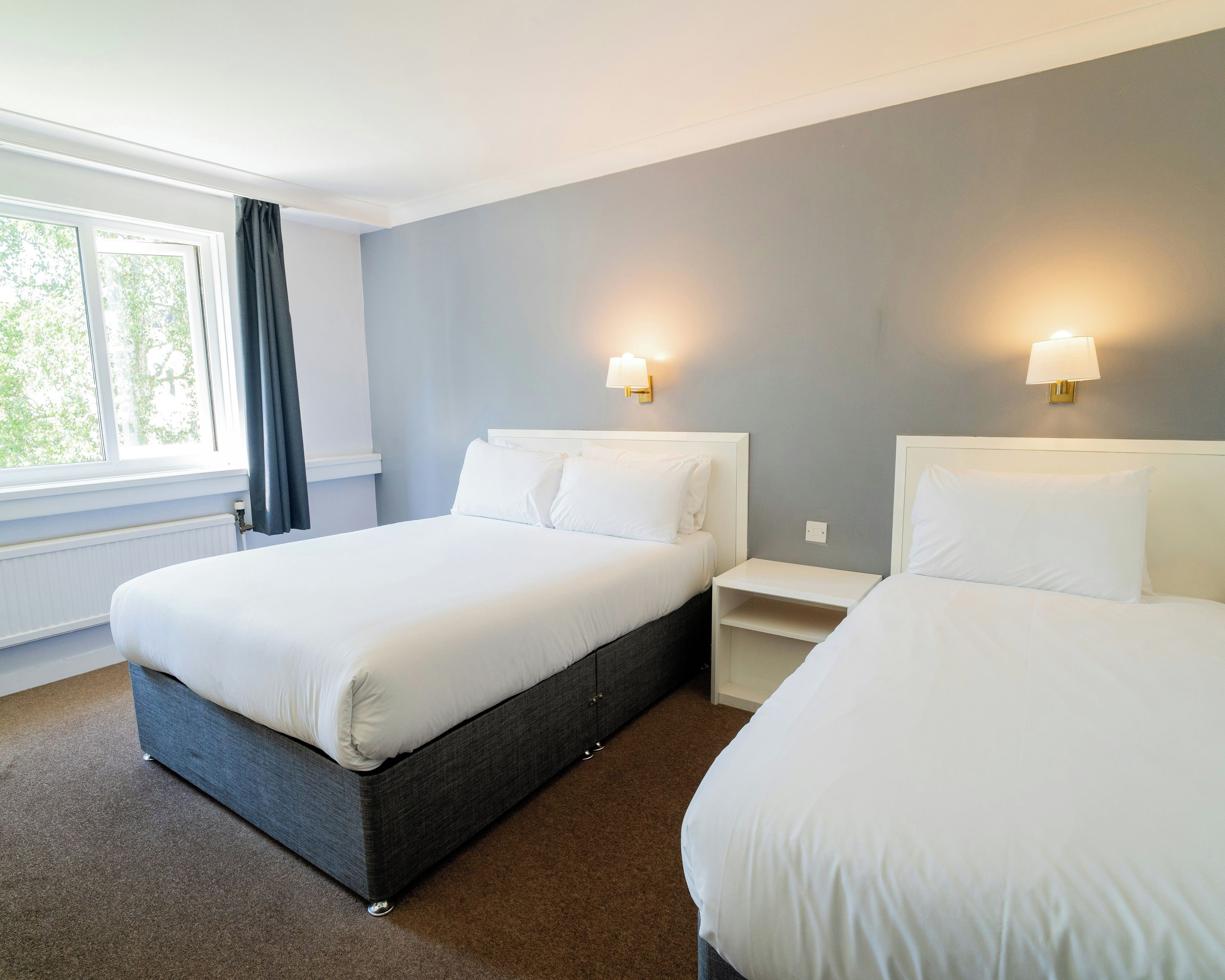 A hotel room with two beds, a window with curtains, and wall-mounted lamps.
