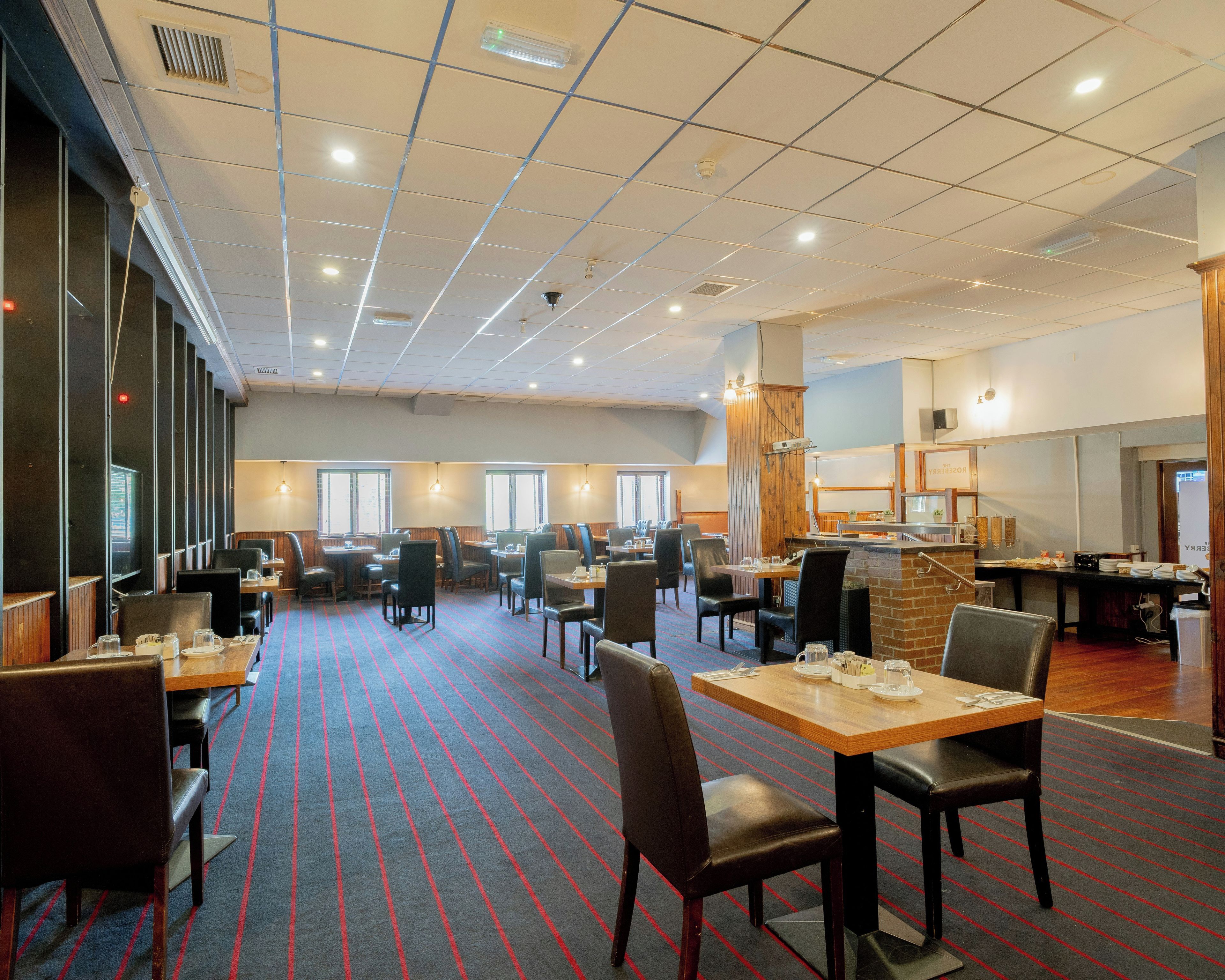 A hotel dining area with tables and chairs, a patterned carpet, and a high ceiling with recessed lighting.
