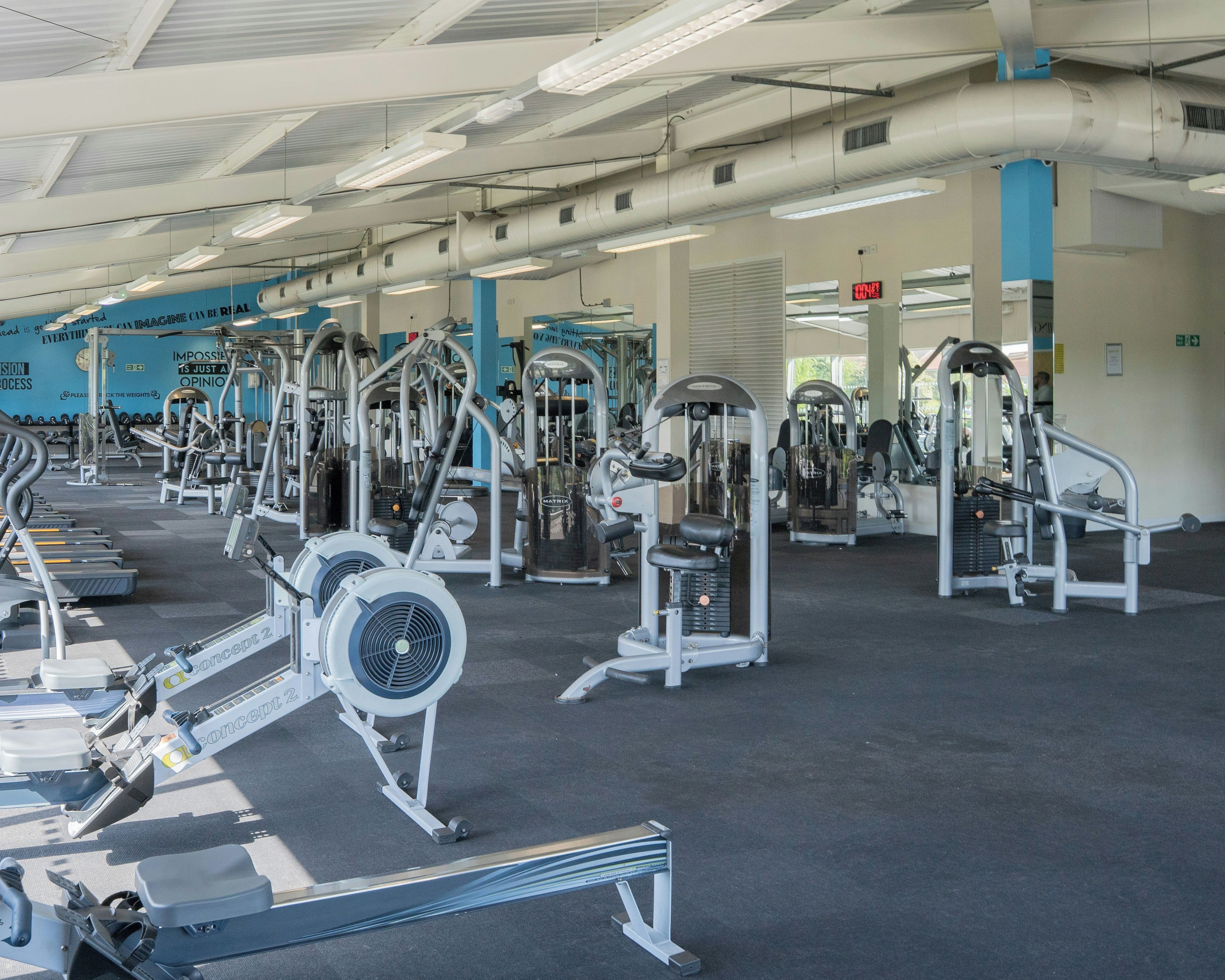 A gym with various exercise machines, a clock, motivational posters, and a digital display.