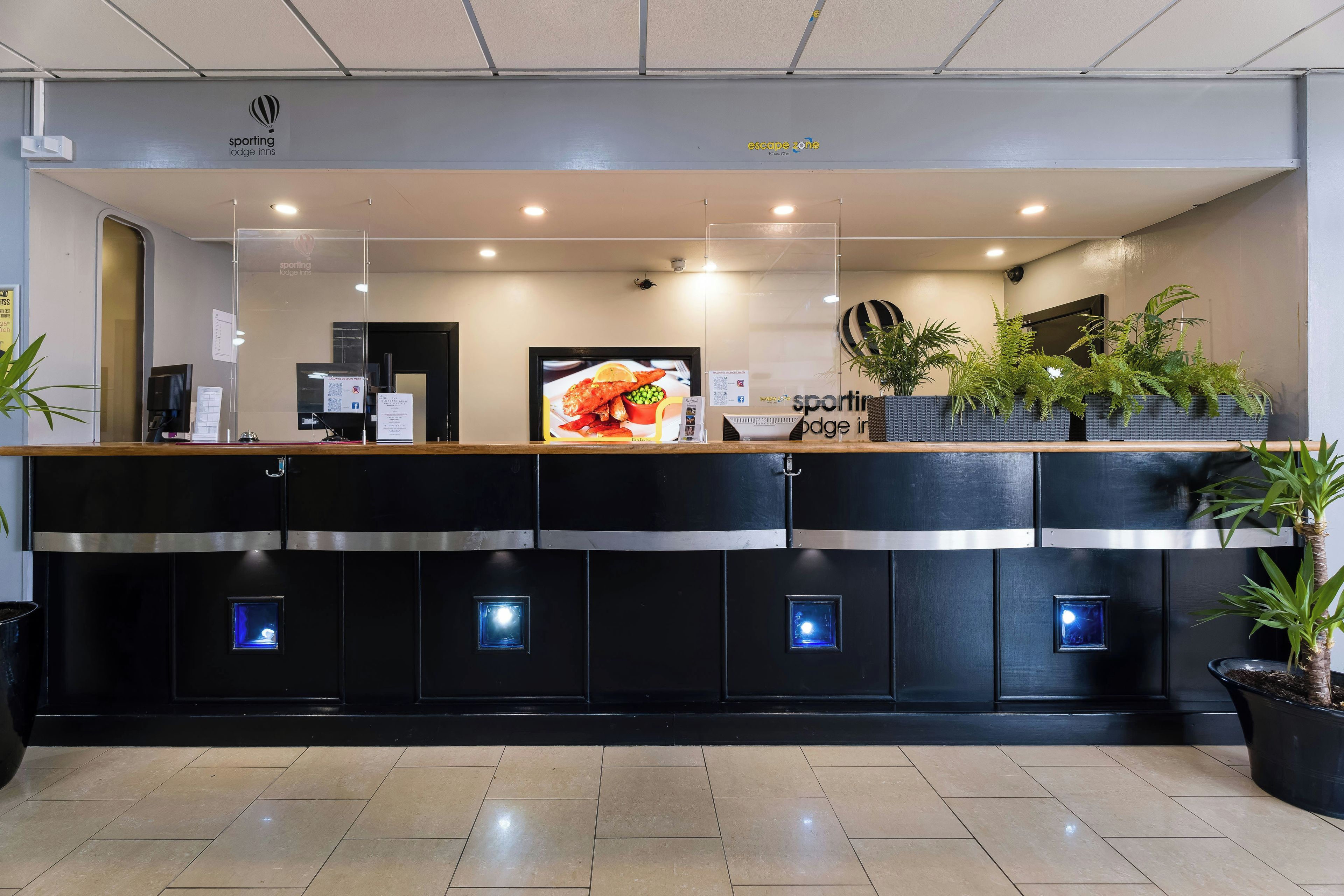 Zona de recepción de un hotel con mostrador, escritorio de recepción y una pantalla grande mostrando una comida.