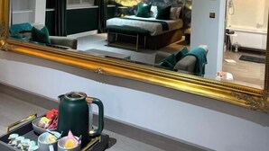 Quarto de hotel com um espelho grande, uma cama com roupa de cama verde, uma mesinha de cabeceira com uma chaleira e chá, e um banheiro visível no reflexo.