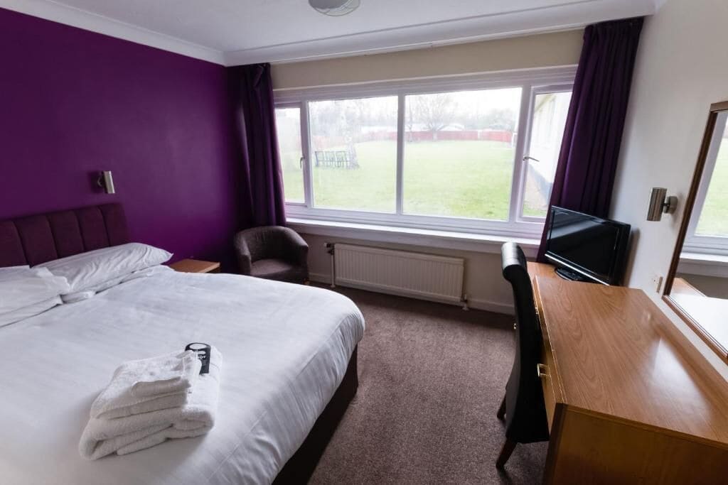 A hotel room with a bed, a desk, a chair, a TV, and a window with a view of a grassy area.