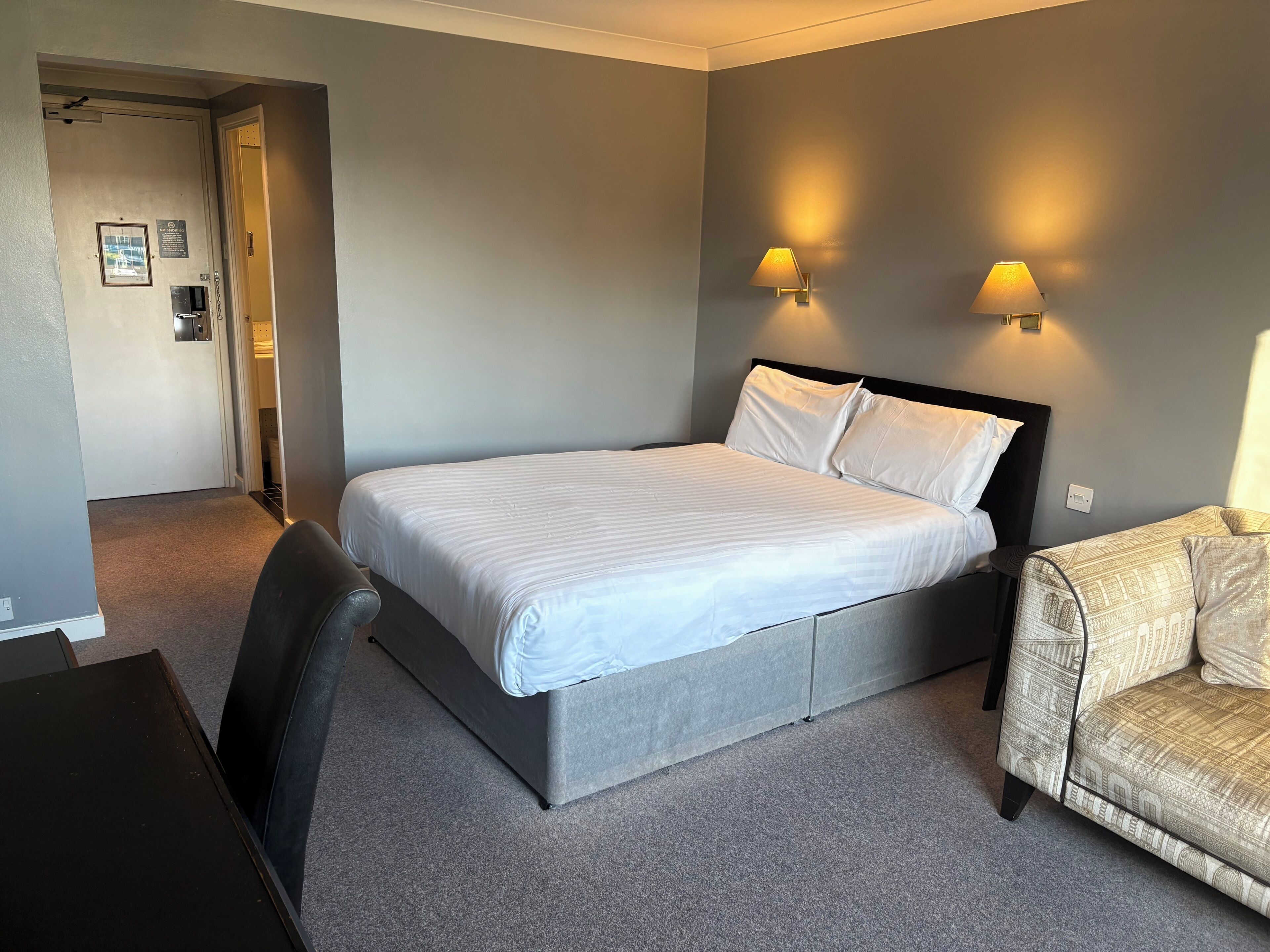 A hotel room with a bed, a chair, a desk, and two wall-mounted lamps.