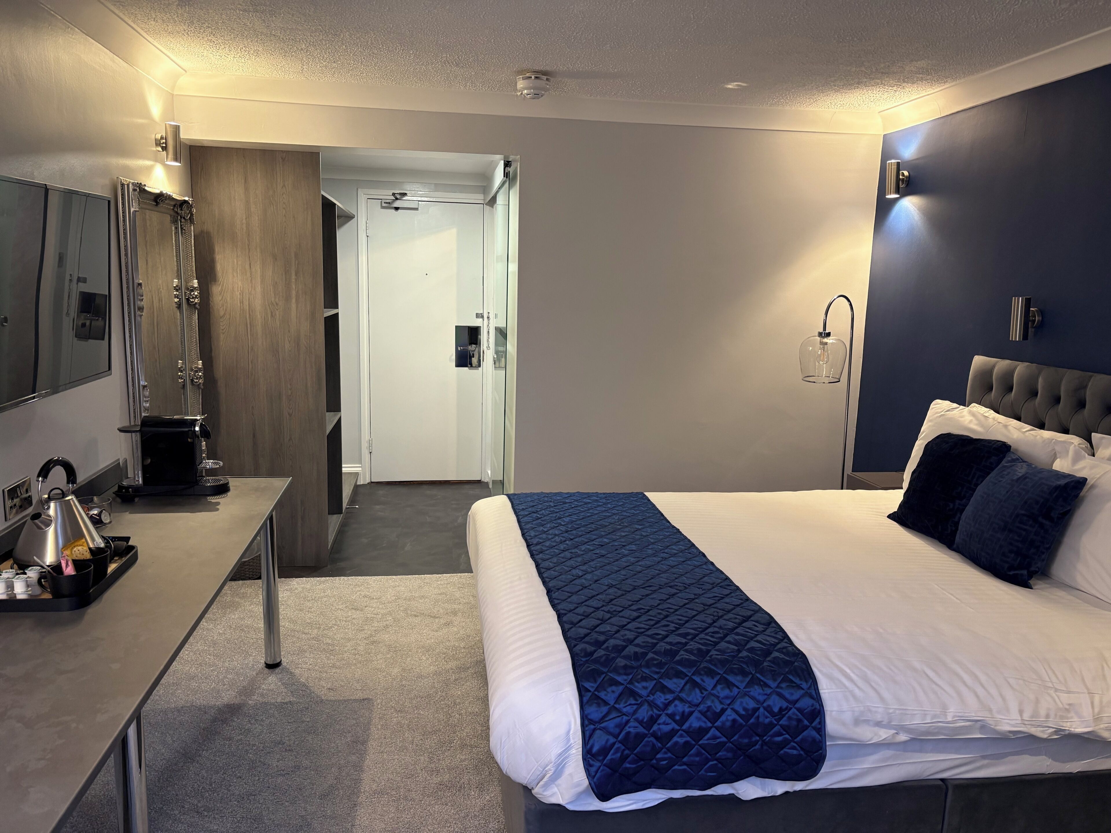 A hotel room with a bed, a desk with a kettle, a mirror, and a wardrobe.