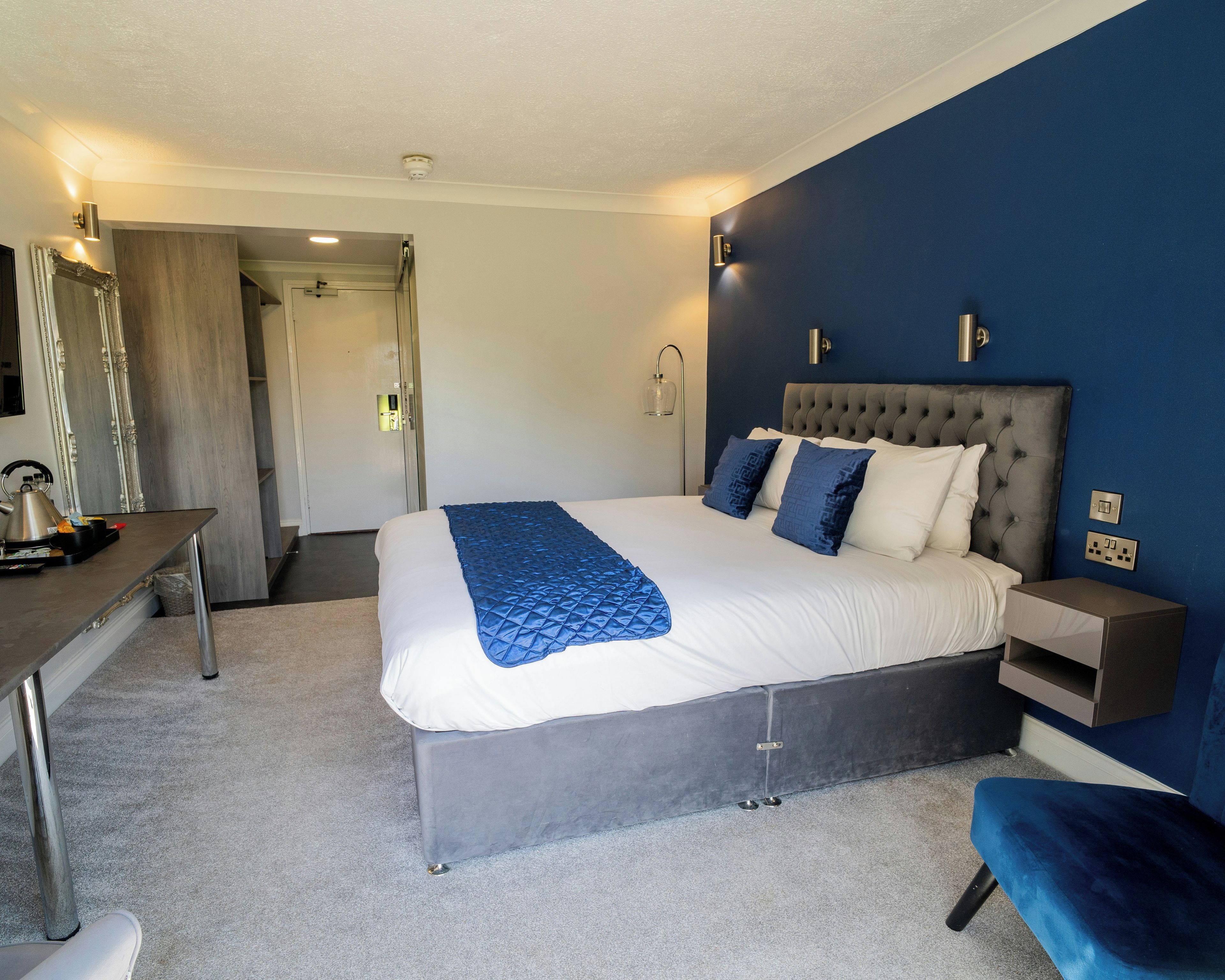 A hotel room with a bed, bedside table, lamp, and a desk with a chair.