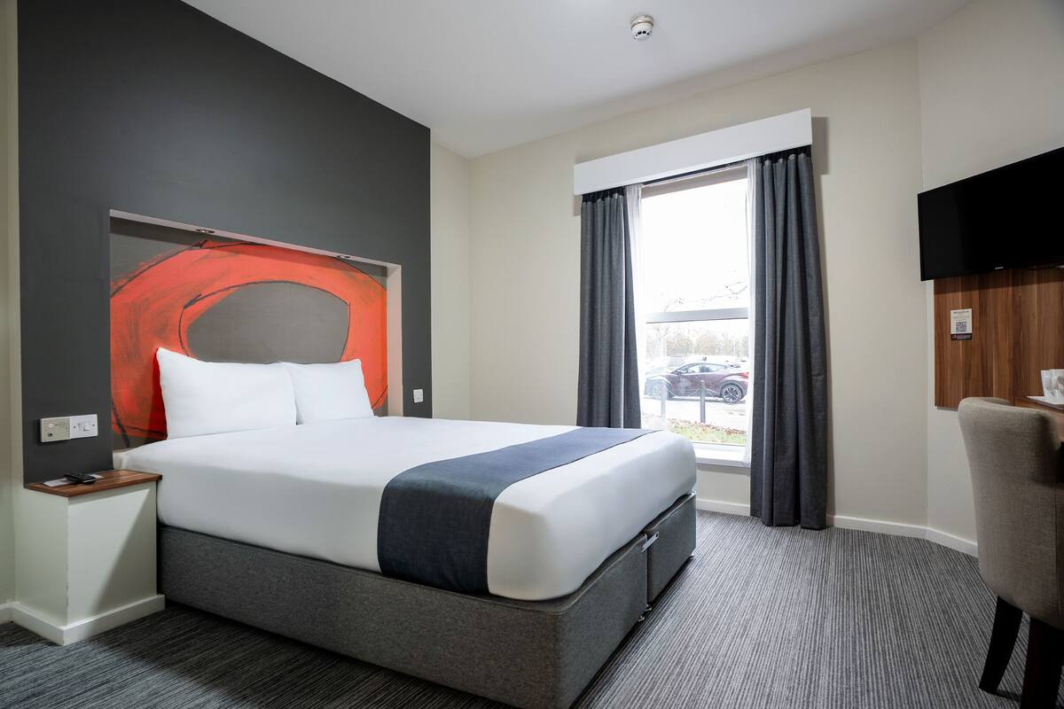 A hotel room with a large bed, a flat-screen TV, a window with curtains, and a view of the outside.