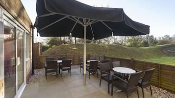 Um pátio externo com mesa e cadeiras, um grande guarda-sol e vista para uma colina gramada.