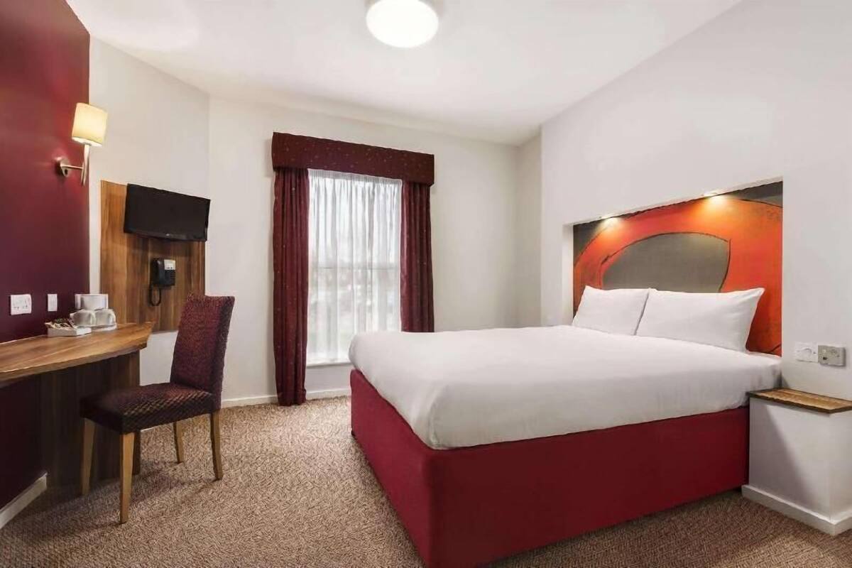 A hotel room with a bed, a desk with a chair, a TV mounted on the wall, and a window with curtains.