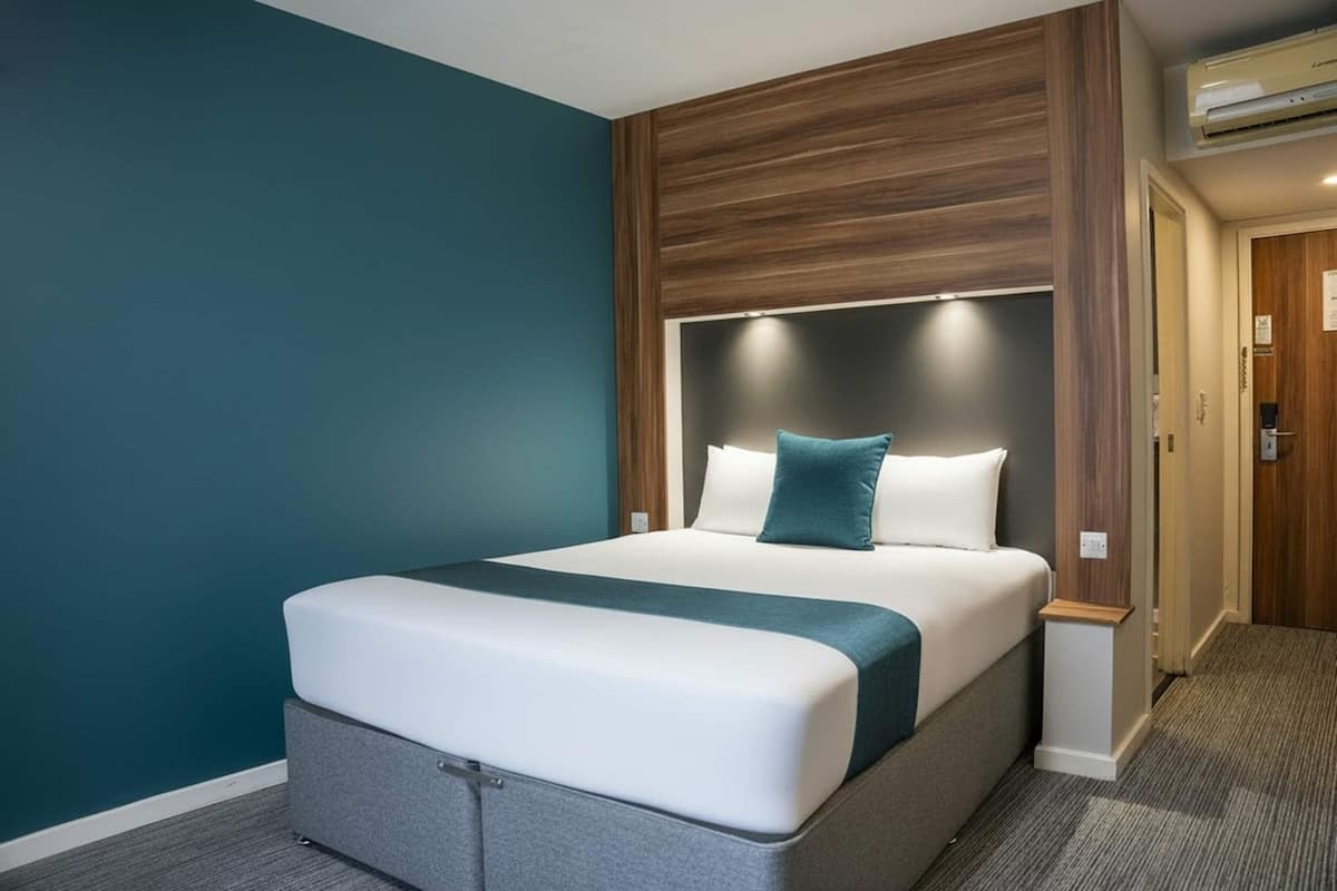 A modern hotel room with a large bed, a wooden headboard, and teal walls.