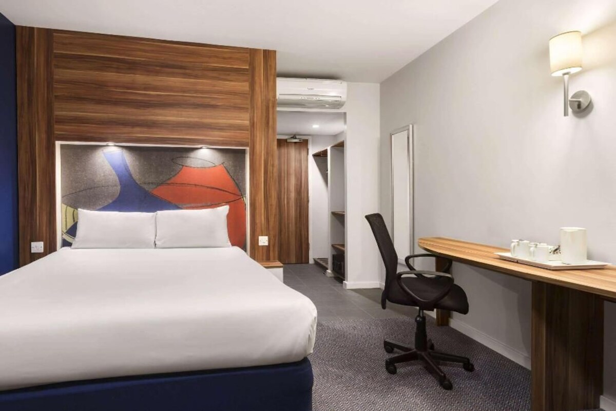 A hotel room with a bed, a desk, a chair, and a lamp.