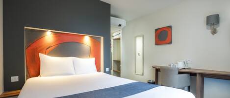 Una habitación de hotel con una cama grande, un escritorio con silla, una lámpara de pared y un cuadro enmarcado.