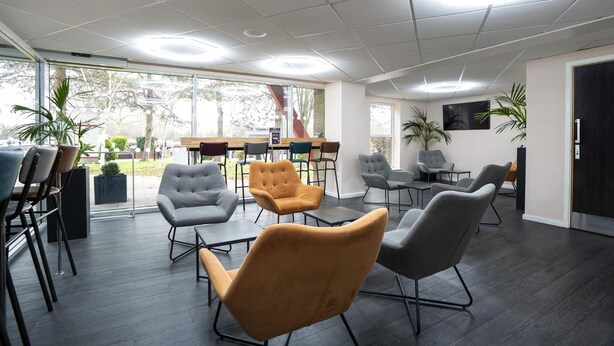 A modern meeting room with dark wooden floors, grey and mustard-colored chairs, and a large window providing a view of the outdoors. - Ramada by Wyndham London Stansted Airport (Bishop's Stortford)