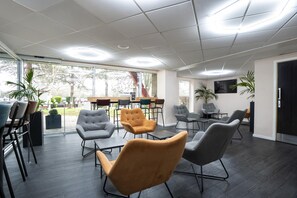 A modern meeting room with dark wooden floors, grey and mustard-colored chairs, and a large window providing a view of the outdoors. - Ramada by Wyndham London Stansted Airport (Bishop's Stortford)