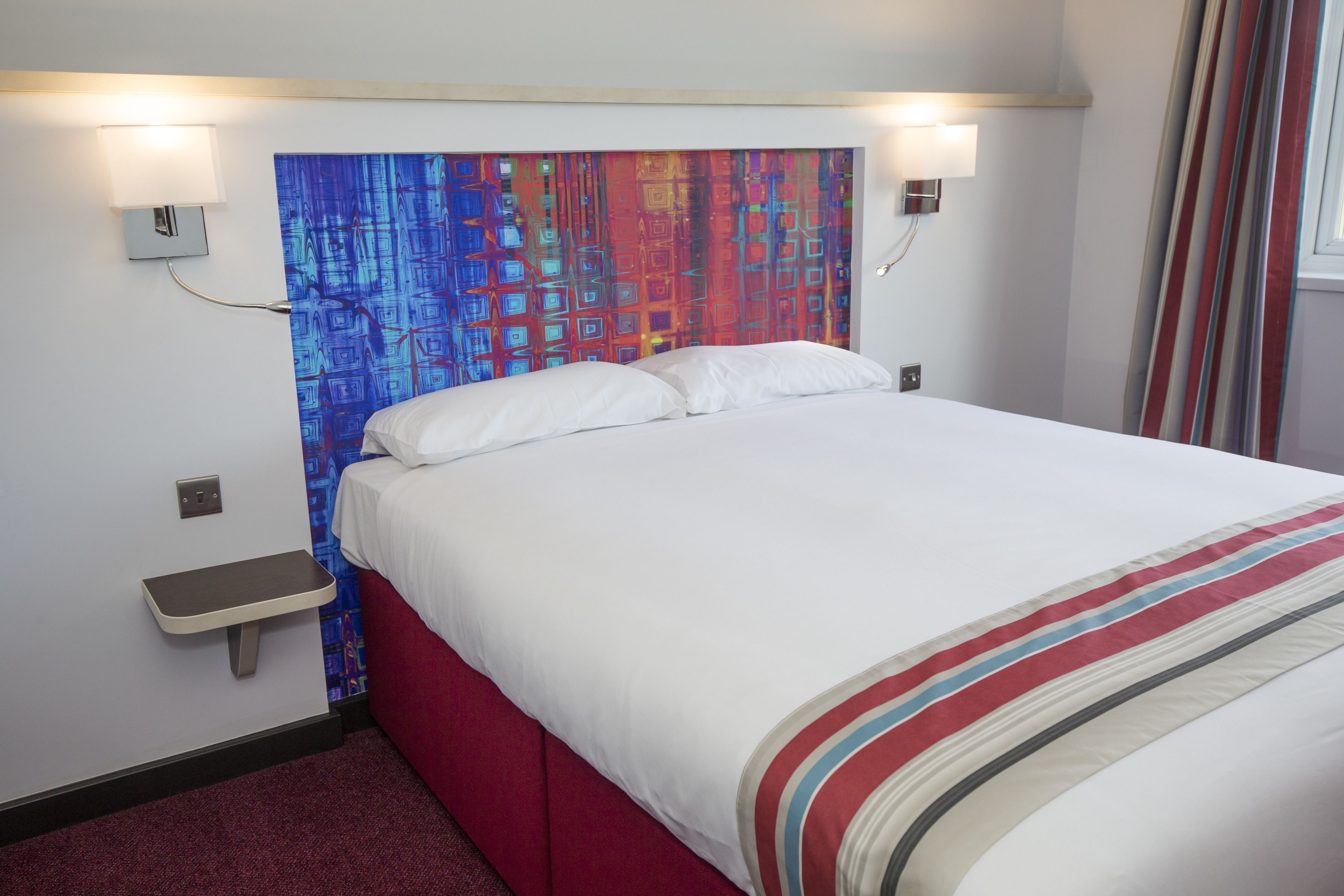 A hotel room with a bed, a nightstand, a wall-mounted lamp, and a colorful abstract painting above the bed.